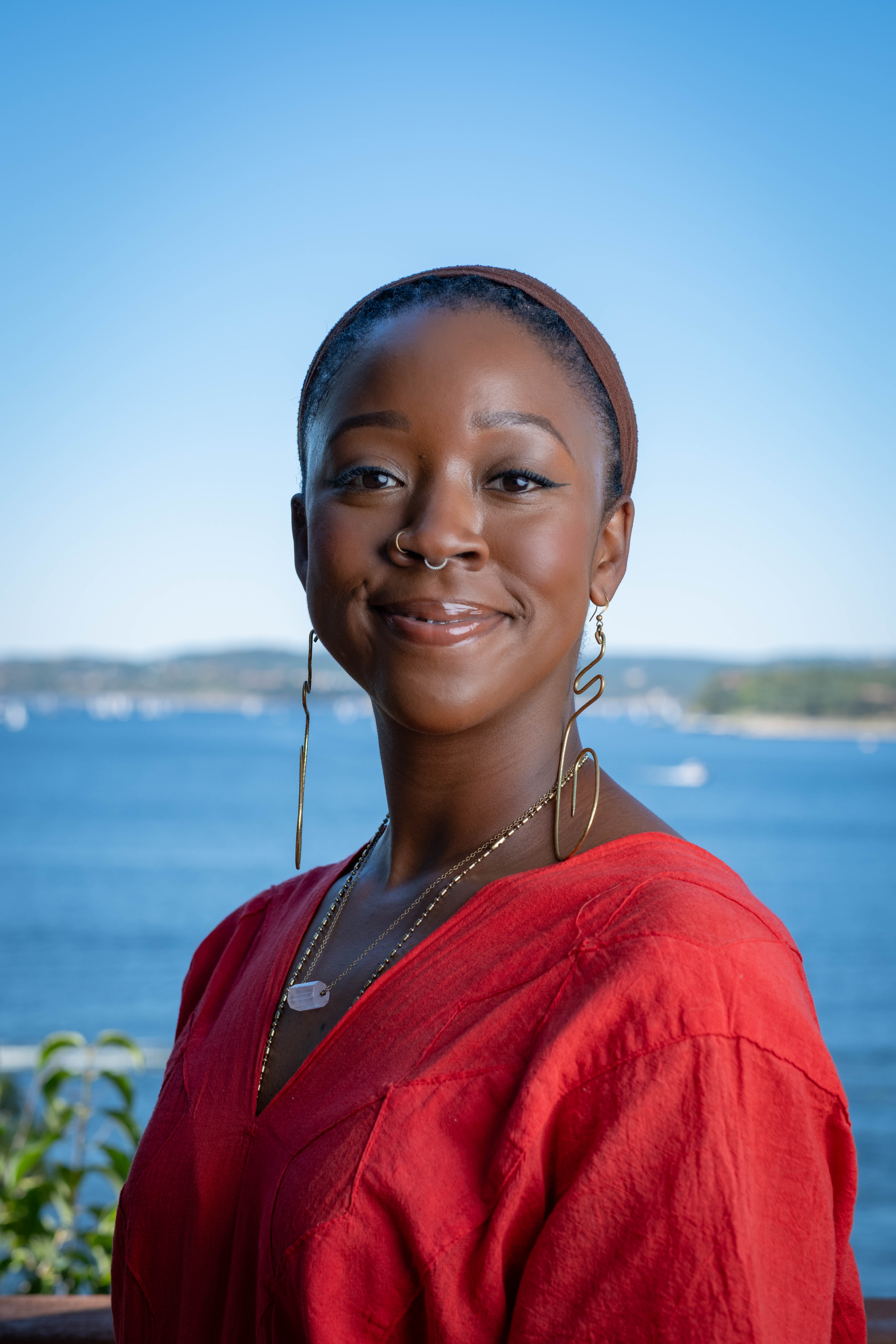 Binti smiles in front of a boat-filled lake during a sunny day. She is wearing a brown headscarf, gold jewelry, and a red huipil.