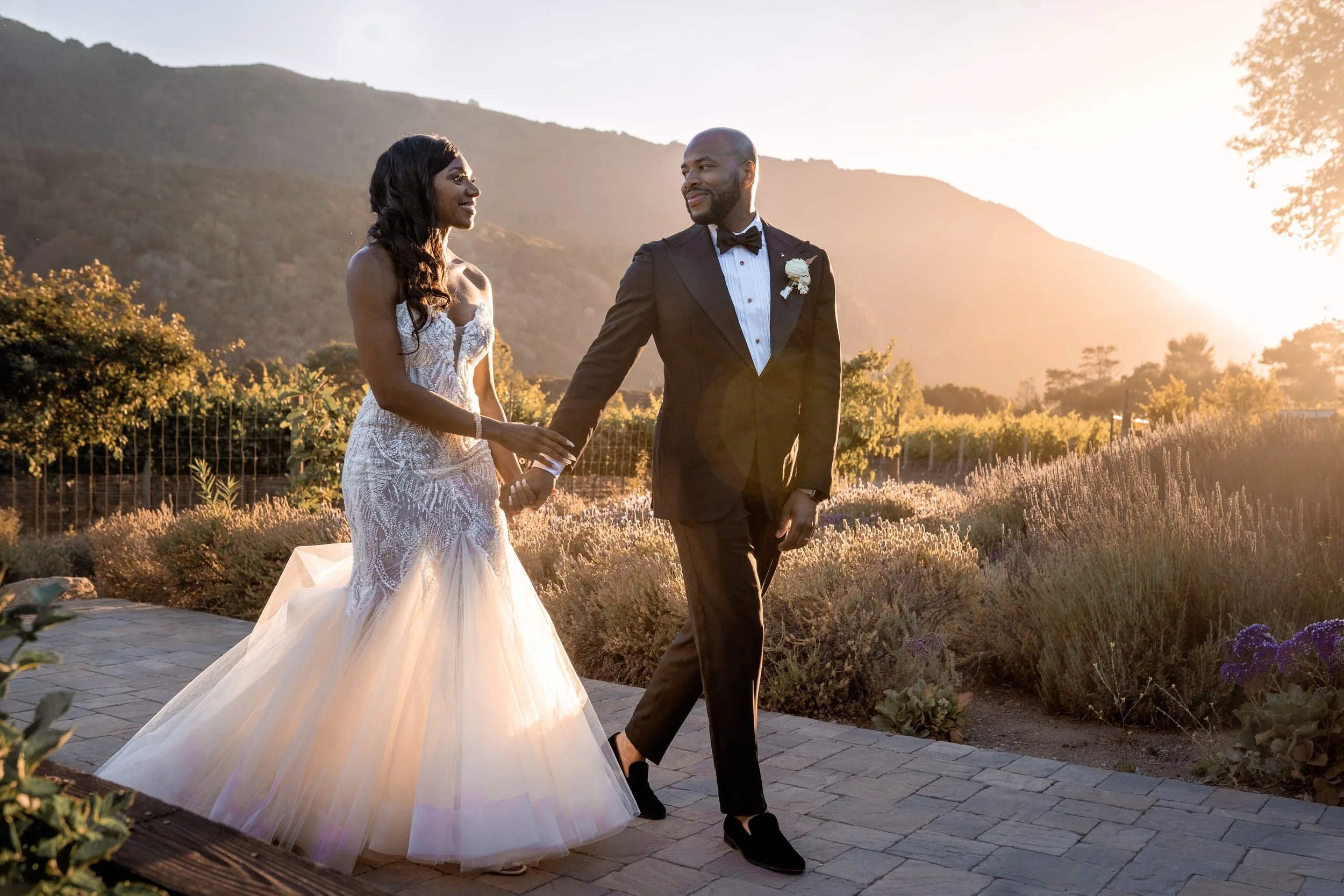 A bride and groom holding hands during their outdoor wedding at sunset, with mountains and trees in the background.