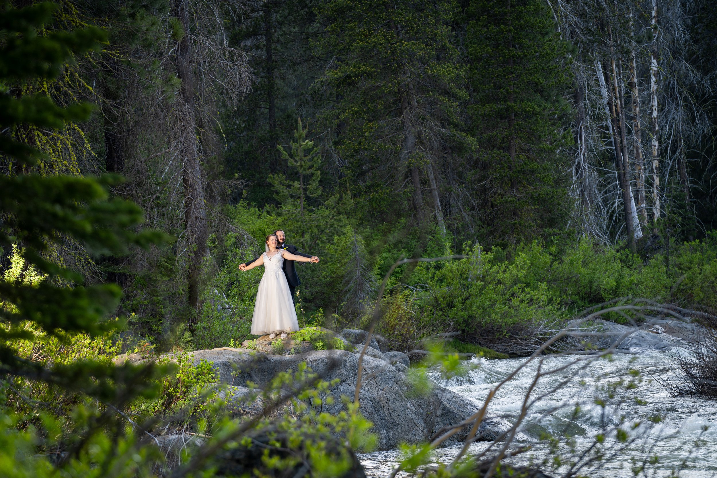 A bride and groom in wedding attire standing on rocks in a forested area near a flowing stream with lush green trees in the background.