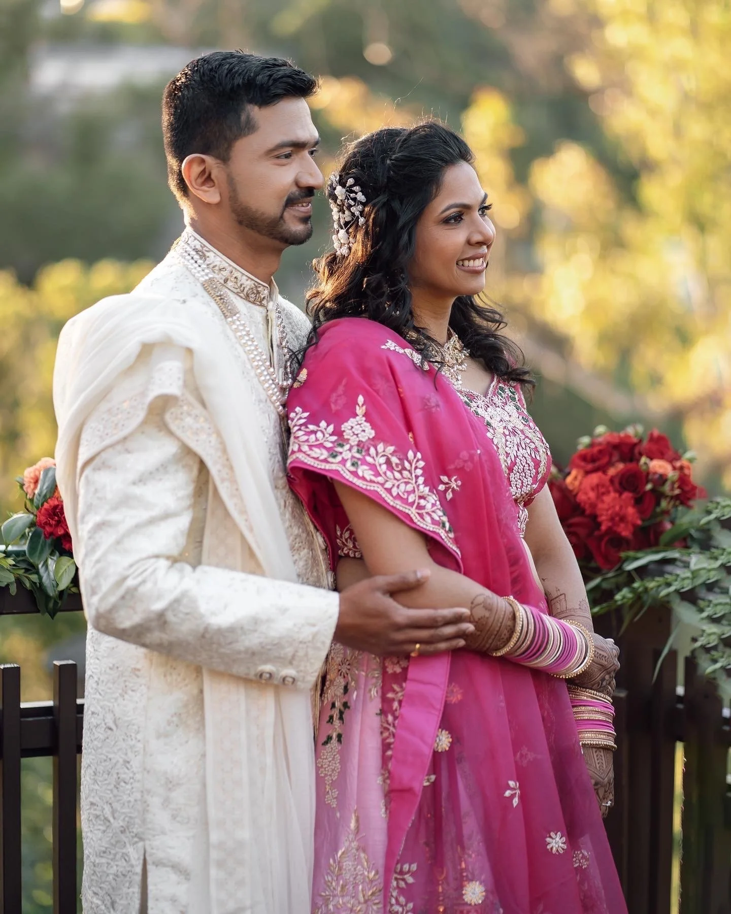 A couple dressed in traditional Indian wedding attire standing outdoors, with the man holding the woman's hand. The scene is illuminated by warm, natural sunlight and decorated with red flowers.