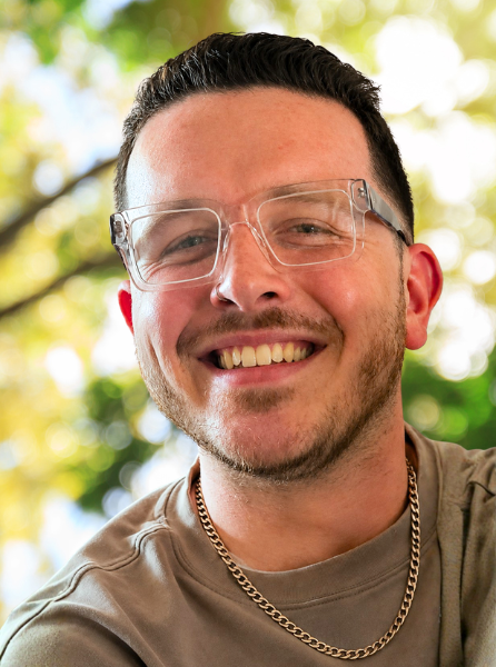A photo of Timothy, a smiling white man with short hair wearing a chain necklace and glasses