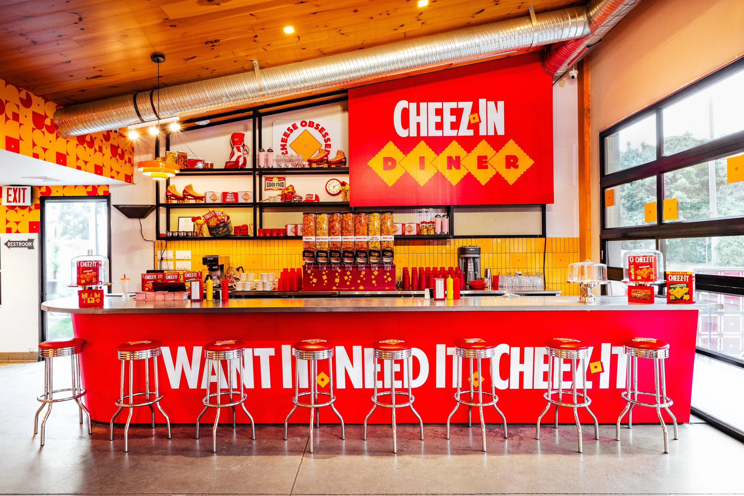 Inside of a Cheez In Diner with a red counter and stools, shelves decorated with Cheez It snacks, and a large Cheez In Diner sign.
