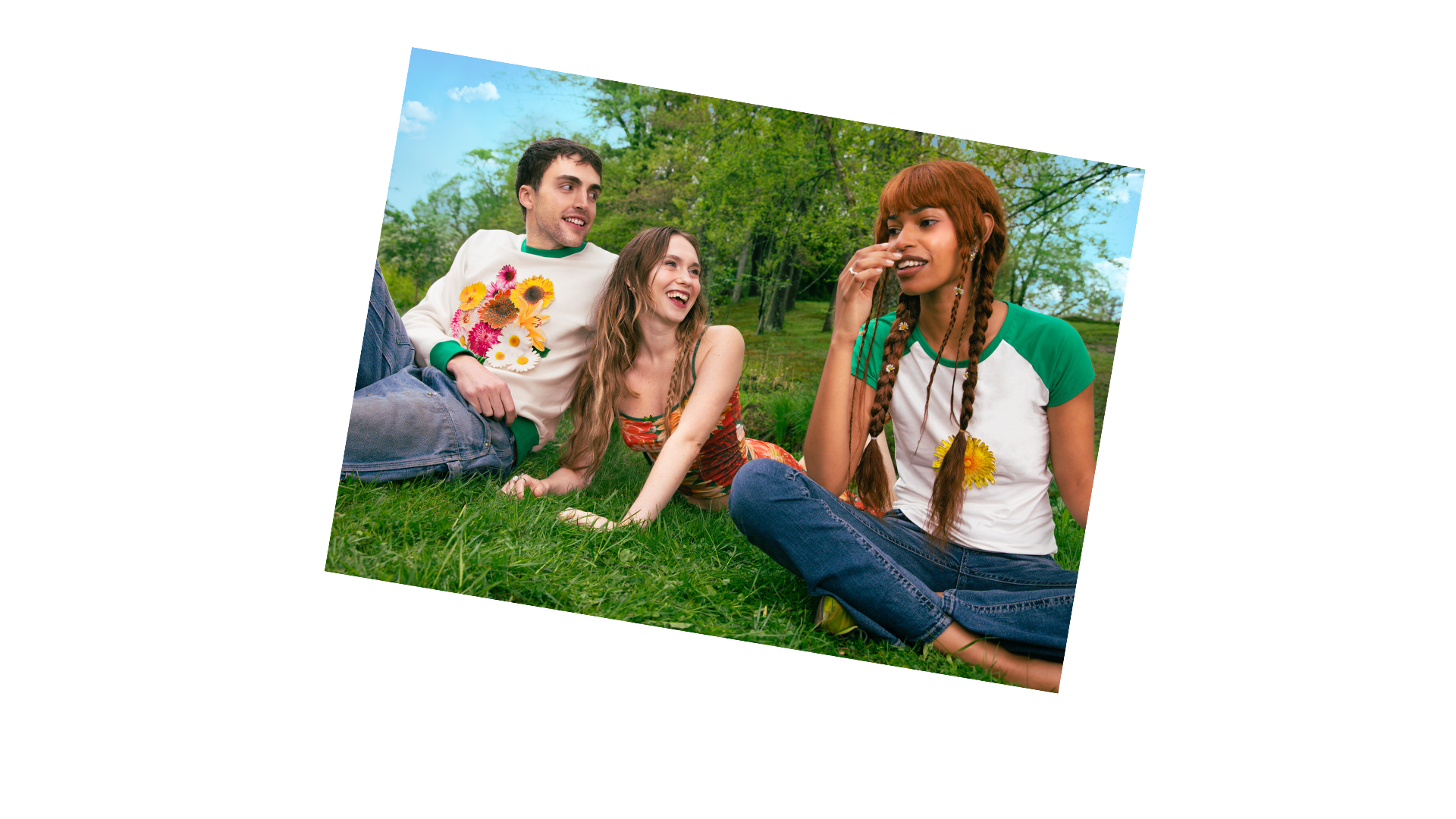 Three friends sitting on grass in a park, smiling and talking. One man and one woman are sitting close together, while another woman with long red hair and braids is sitting on the grass nearby, wearing a t-shirt with flowers, green and white. Trees and blue sky are in the background.