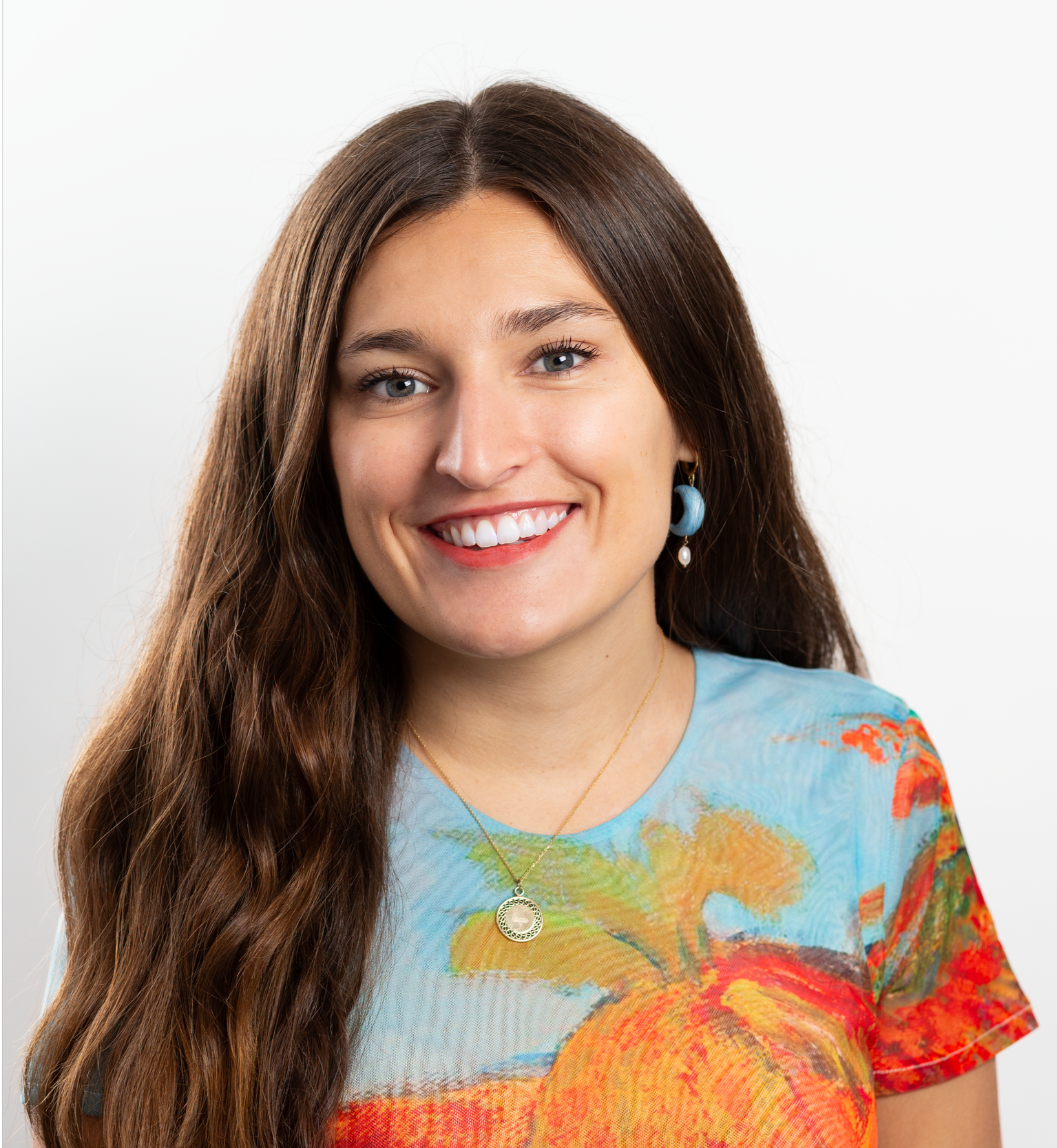 A young woman with long brown hair, light skin, smiling, wearing blue earrings and a colorful shirt with a tropical print, standing against a plain white background.