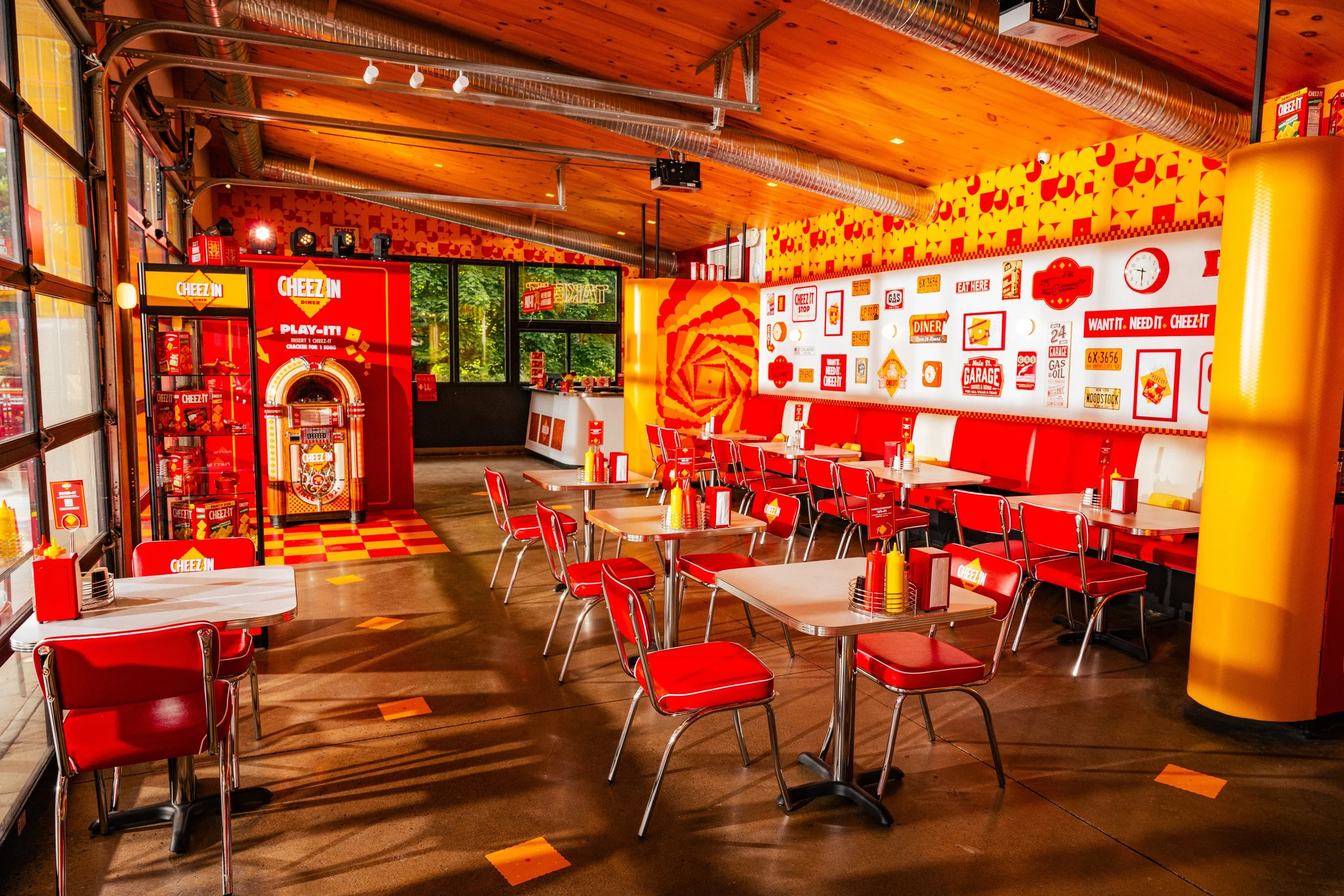 Interior of a Cheez-In themed diner with red and yellow decor, red chairs, tables with condiments, and wall decorations featuring Cheez-In branding and vintage signs.