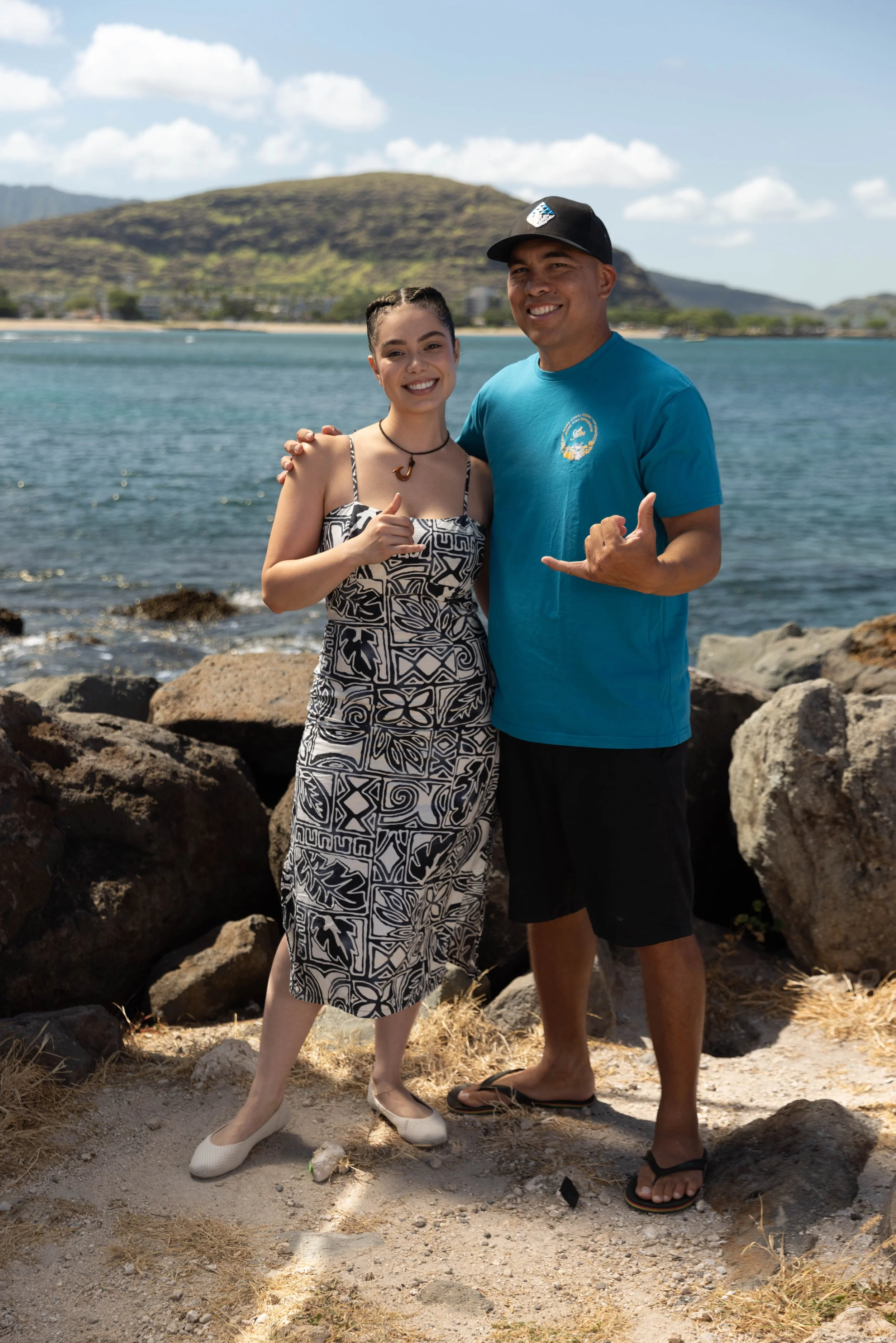 Auli'i Cravalho and a man smiling and making the shaka hand sign, standing on rocks and sandy ground near the water with a hill and cloudy sky in the background.