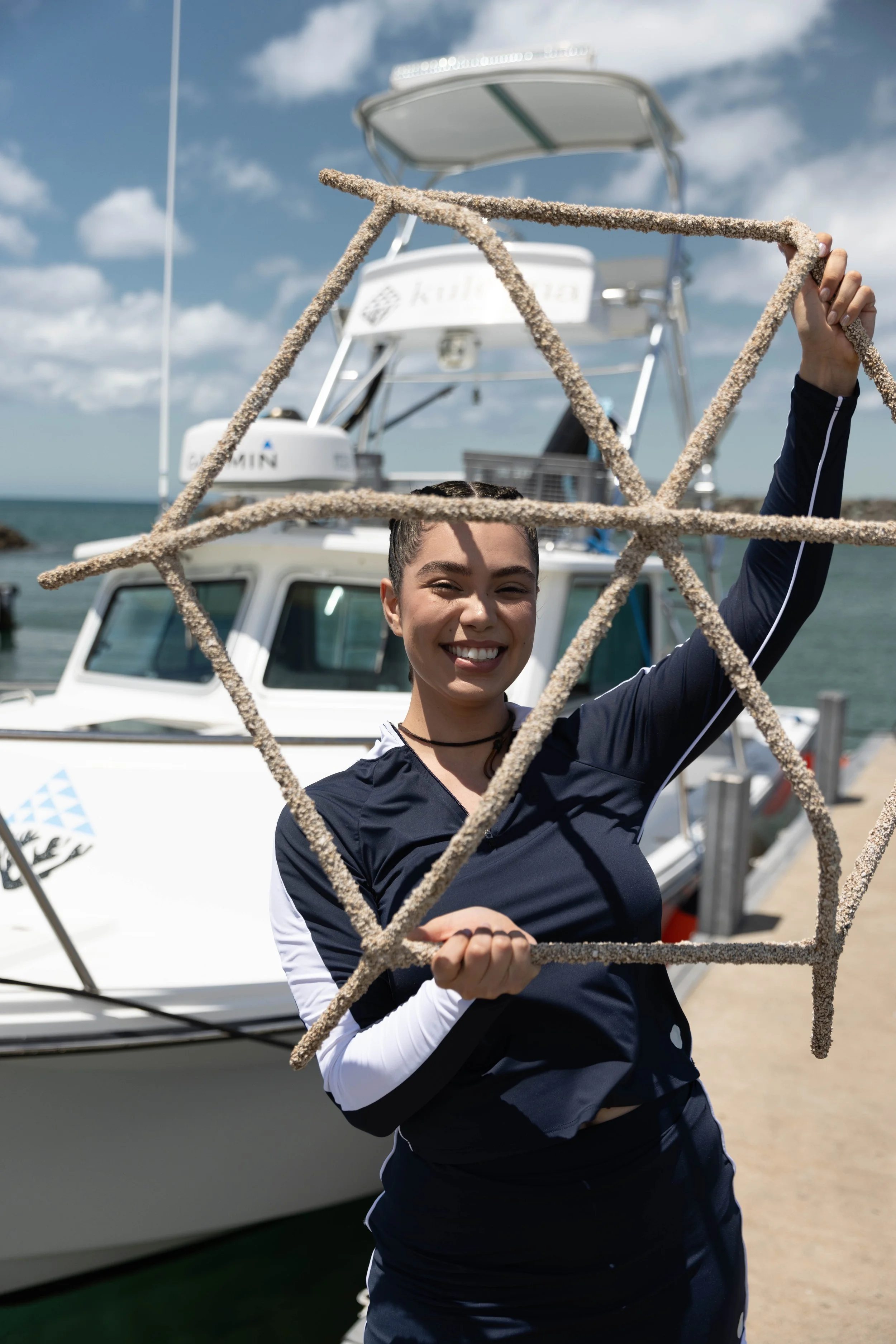 Auli'i Cravalho smiling and holding a marine rope in front of a dock with a boat and blue sky with clouds in the background.