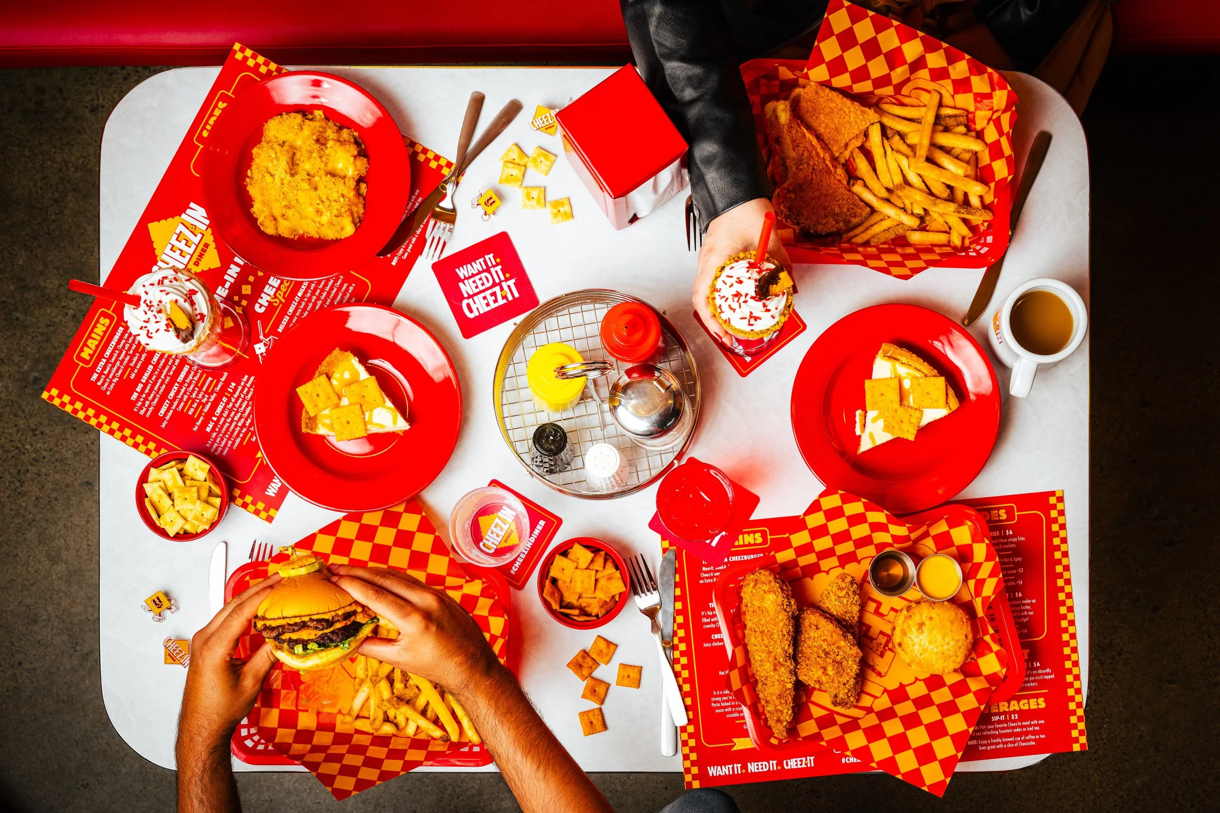 Overhead view of a fast food meal on a table, including a cheeseburger, fries, fried chicken, a milkshake, and a cup of coffee. The table is set with red and yellow checkered paper and red plates with Cheez-It branding.