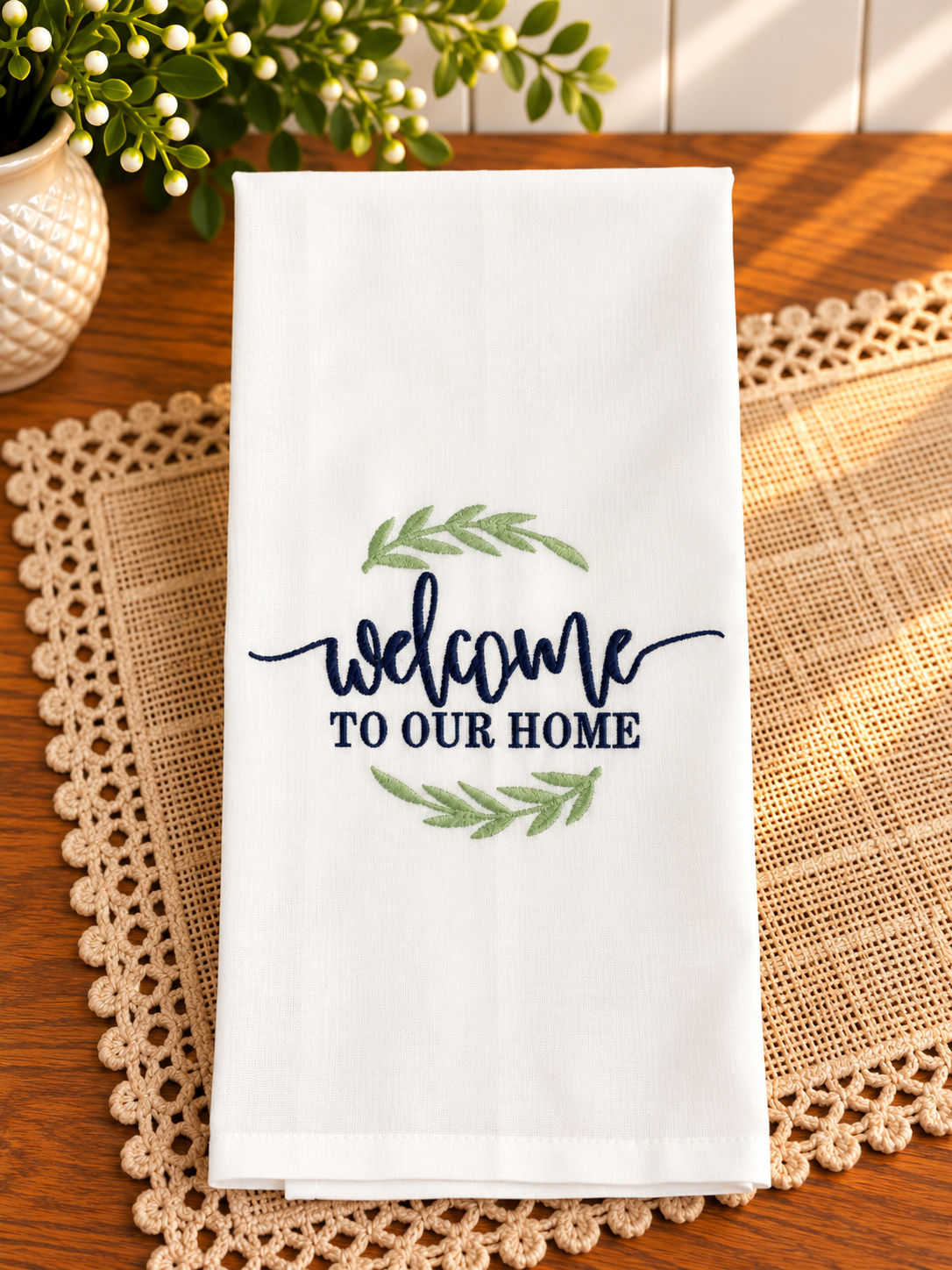 Ivory cotton tea towel with Welcome to Our Home embroidered in navy blue thread, accented by sage green leaves and displayed on a warm wooden table.