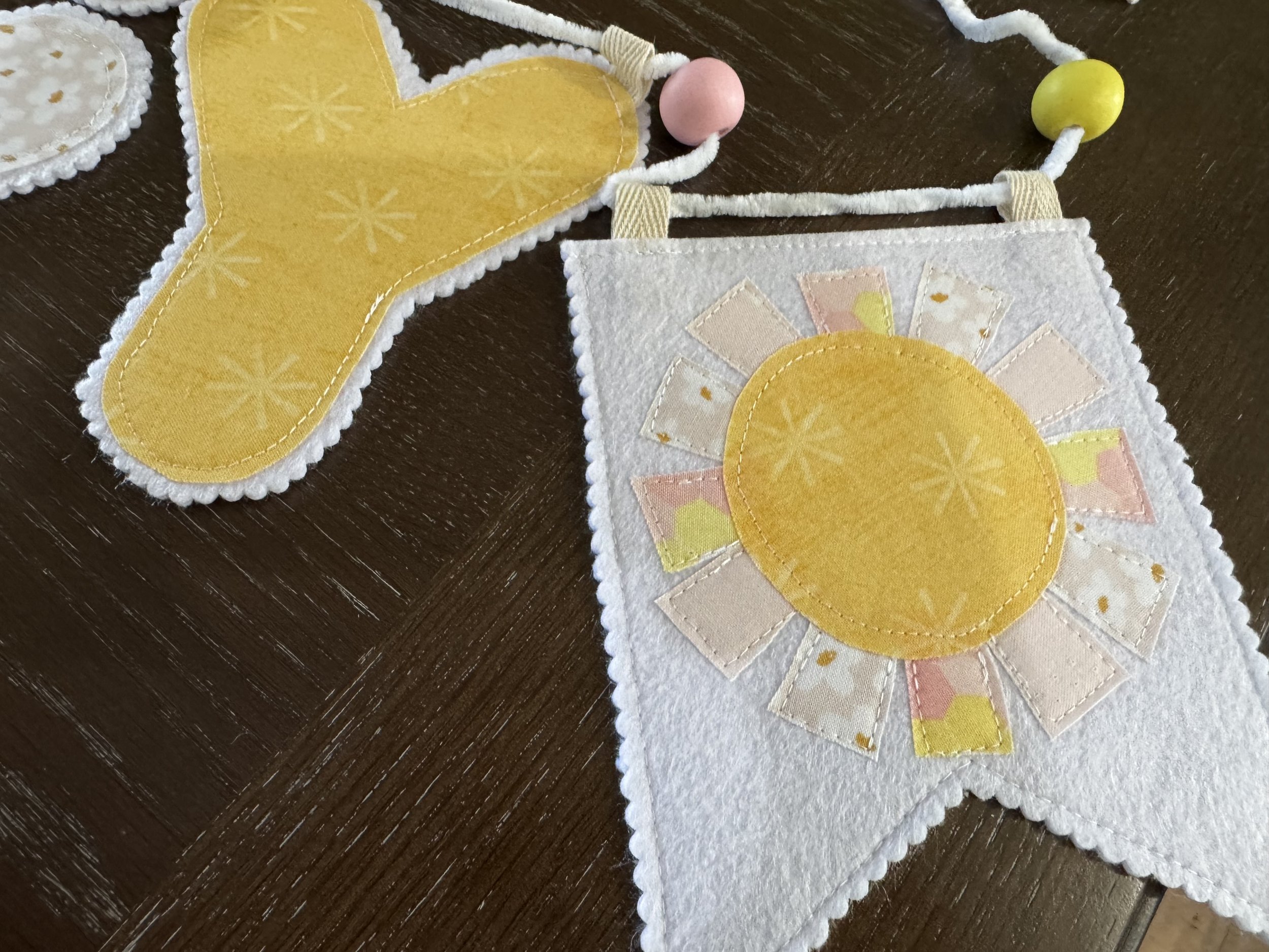 Handmade sunshine fabric appliqué pennant with yellow cotton fabric, pastel sun rays and white felt backing for a personalized nursery garland.