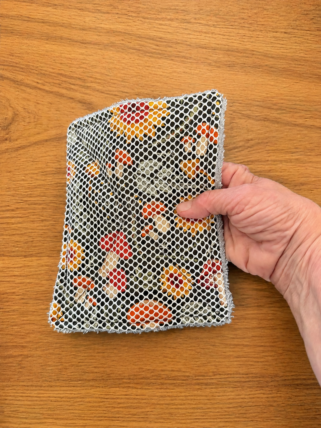 Hand holding a handmade oversized mushroom kitchen scrubbie with white vinyl mesh on a warm wood background