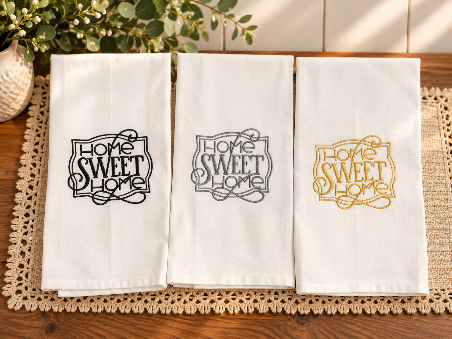 Three ivory cotton tea towels featuring Home Sweet Home embroidery in navy blue, slate gray, and champagne gold, styled on a warm wooden table.