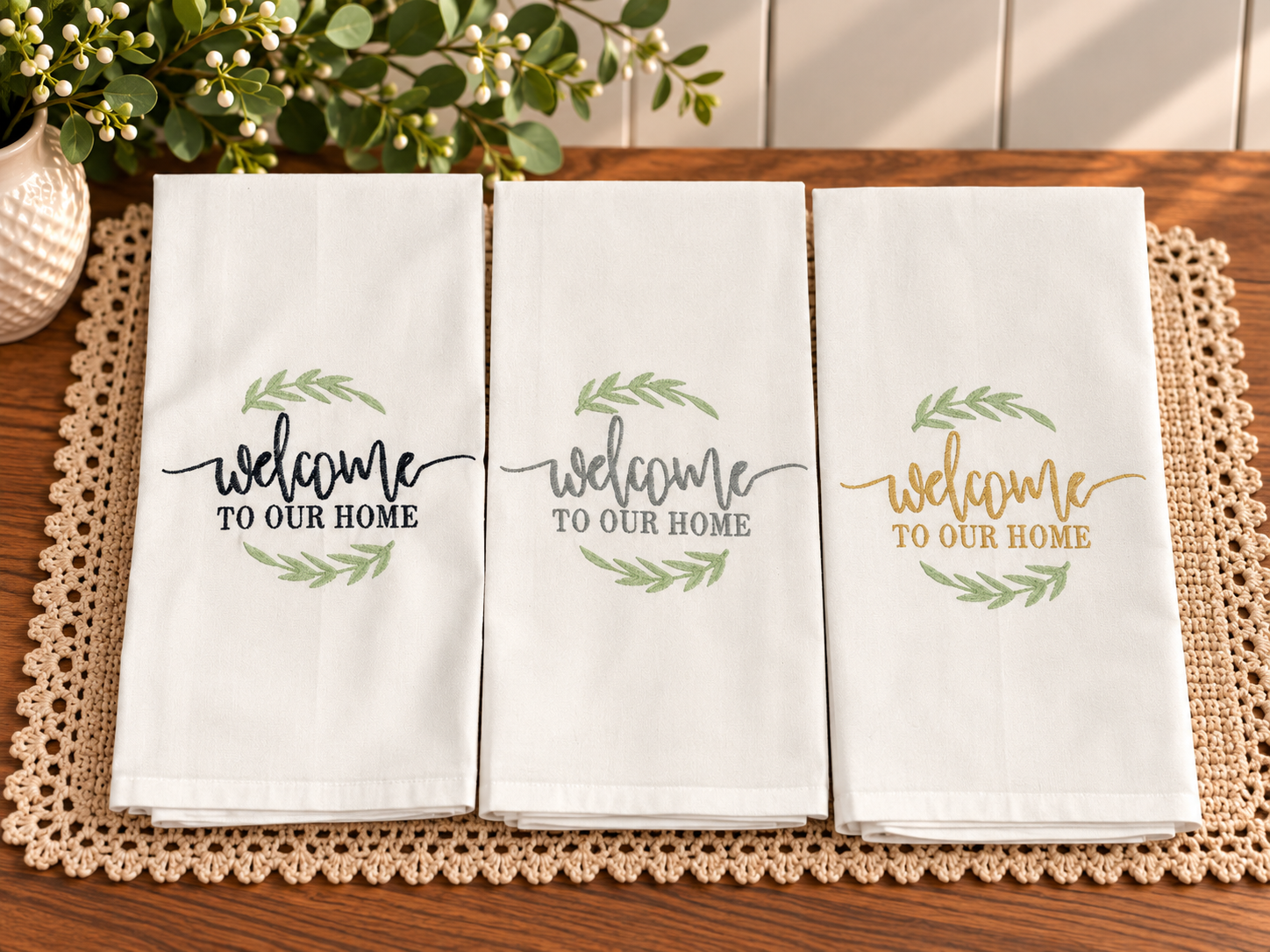 Three ivory cotton tea towels embroidered with Welcome to Our Home lettering in navy blue, slate gray, and champagne gold, accented with sage green leaves and styled on a wooden table.
