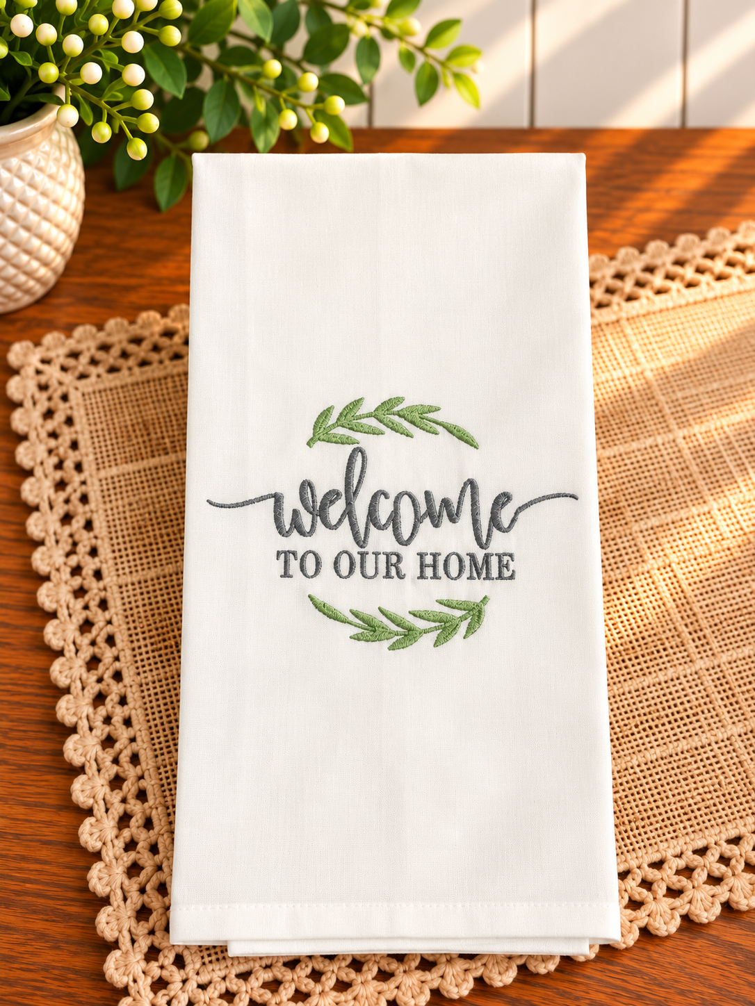 Ivory cotton tea towel with Welcome to Our Home embroidered in slate gray thread, framed by soft sage green leafy accents on a warm wooden table.