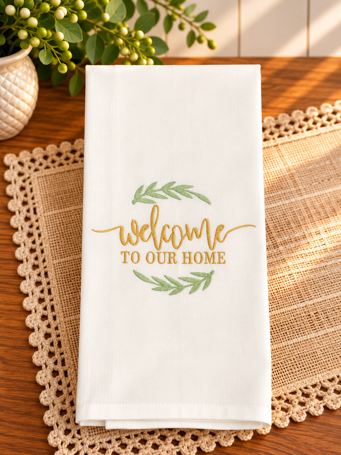 Ivory cotton tea towel featuring Welcome to Our Home embroidery in champagne gold thread with sage green leafy accents, styled on a warm wooden table.