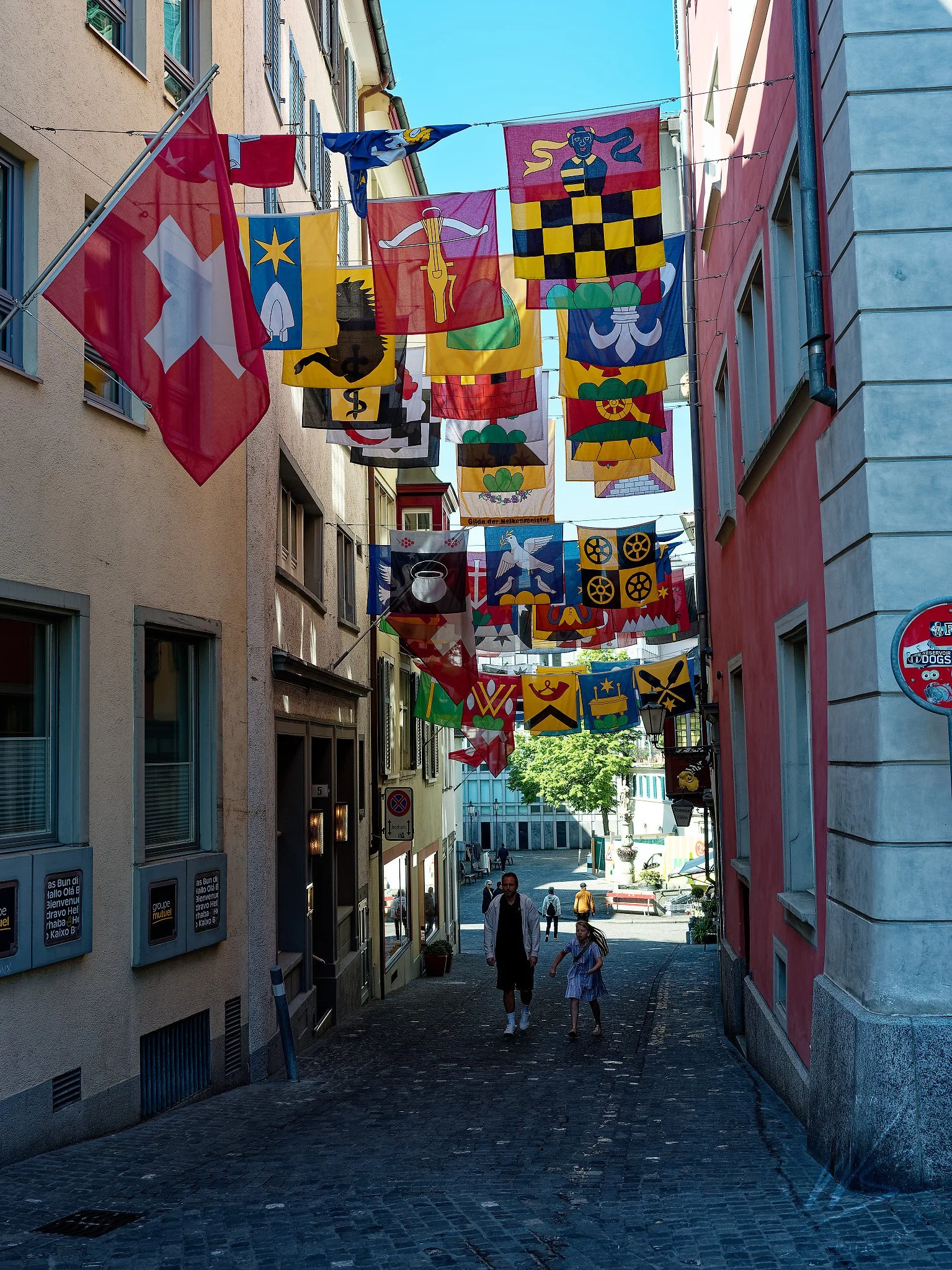 Guild flags (Zunftfahnen) strung across a narrow cobblestone alley in Zurich Old Town, Zurich, Switzerland. The colourful heraldic banners represent Zurich's historic guilds (Zünfte) — the powerful medieval trade associations that governed the city f