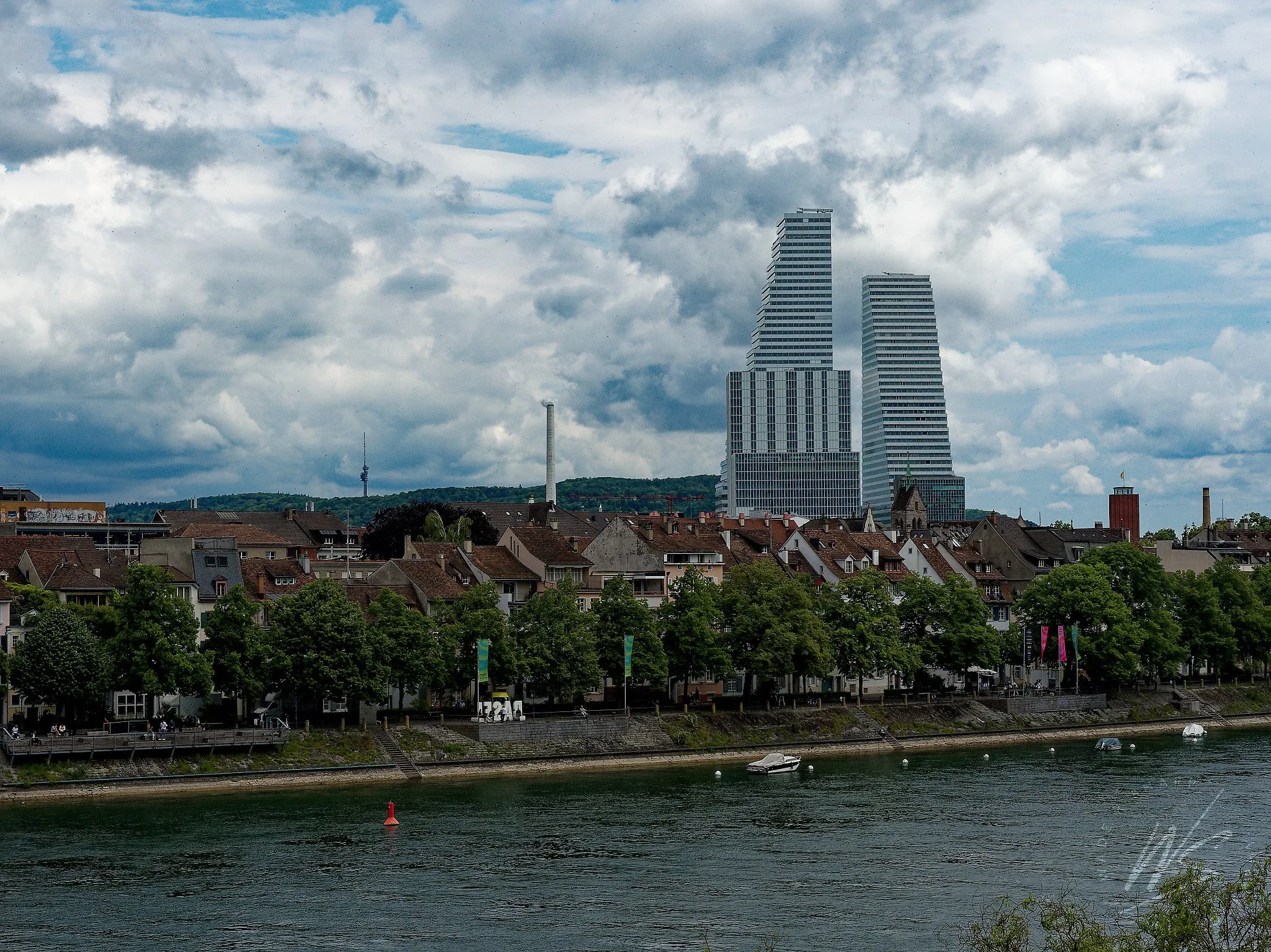 The Rhine River and Kleinbasel riverfront, Basel, Switzerland, with the two landmark Roche Towers — designed by Herzog & de Meuron on the F. Hoffmann-La Roche pharmaceutical campus — rising above the city rooftops. The St. Chrischona transmission tow