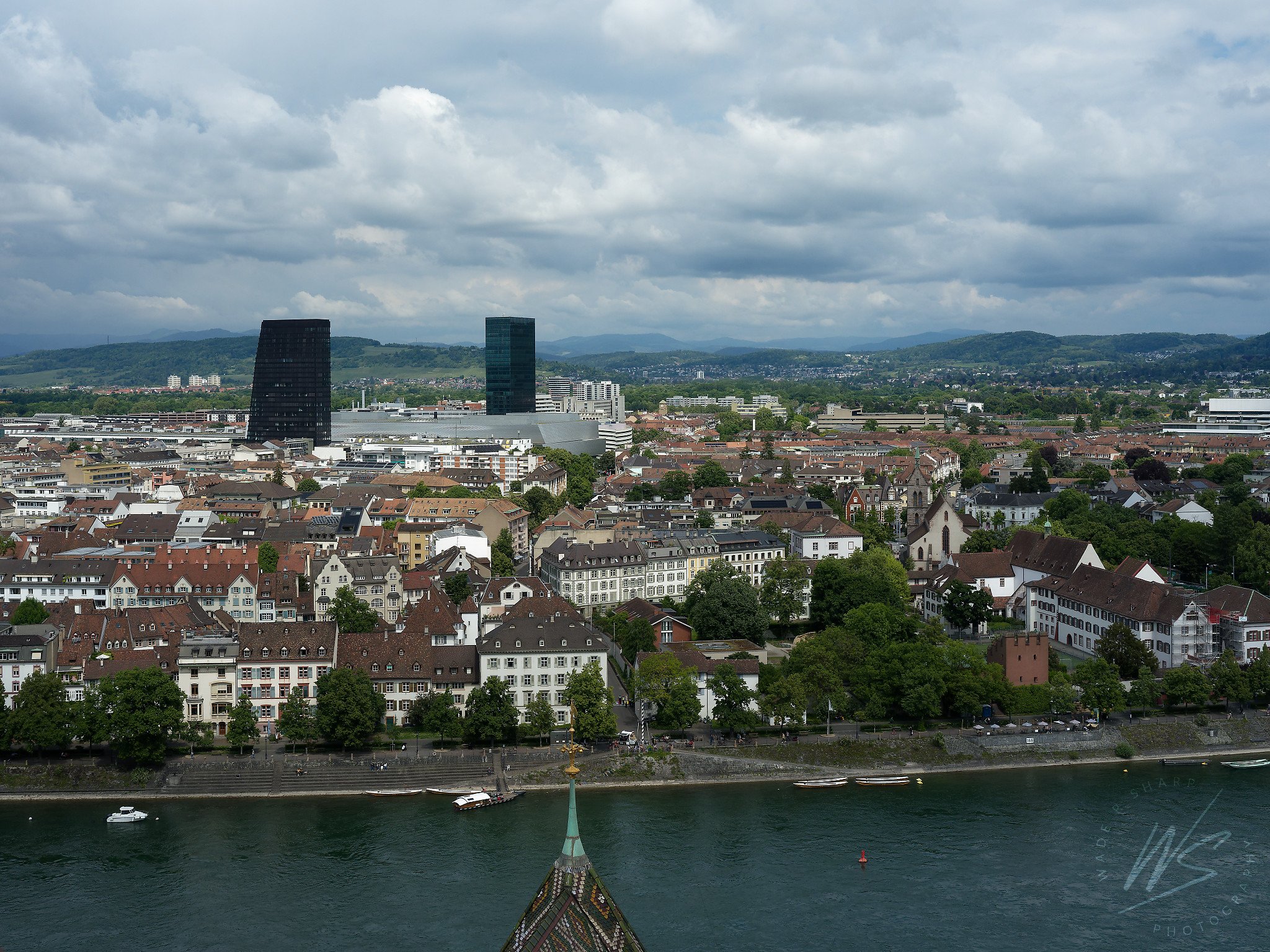 View from the tower of the Basler Münster (Basel Cathedral) looking north across the Rhine River toward Kleinbasel and the Novartis pharmaceutical campus, Basel, Switzerland. The tip of the Münster's iconic diamond-patterned glazed tile roof is visib