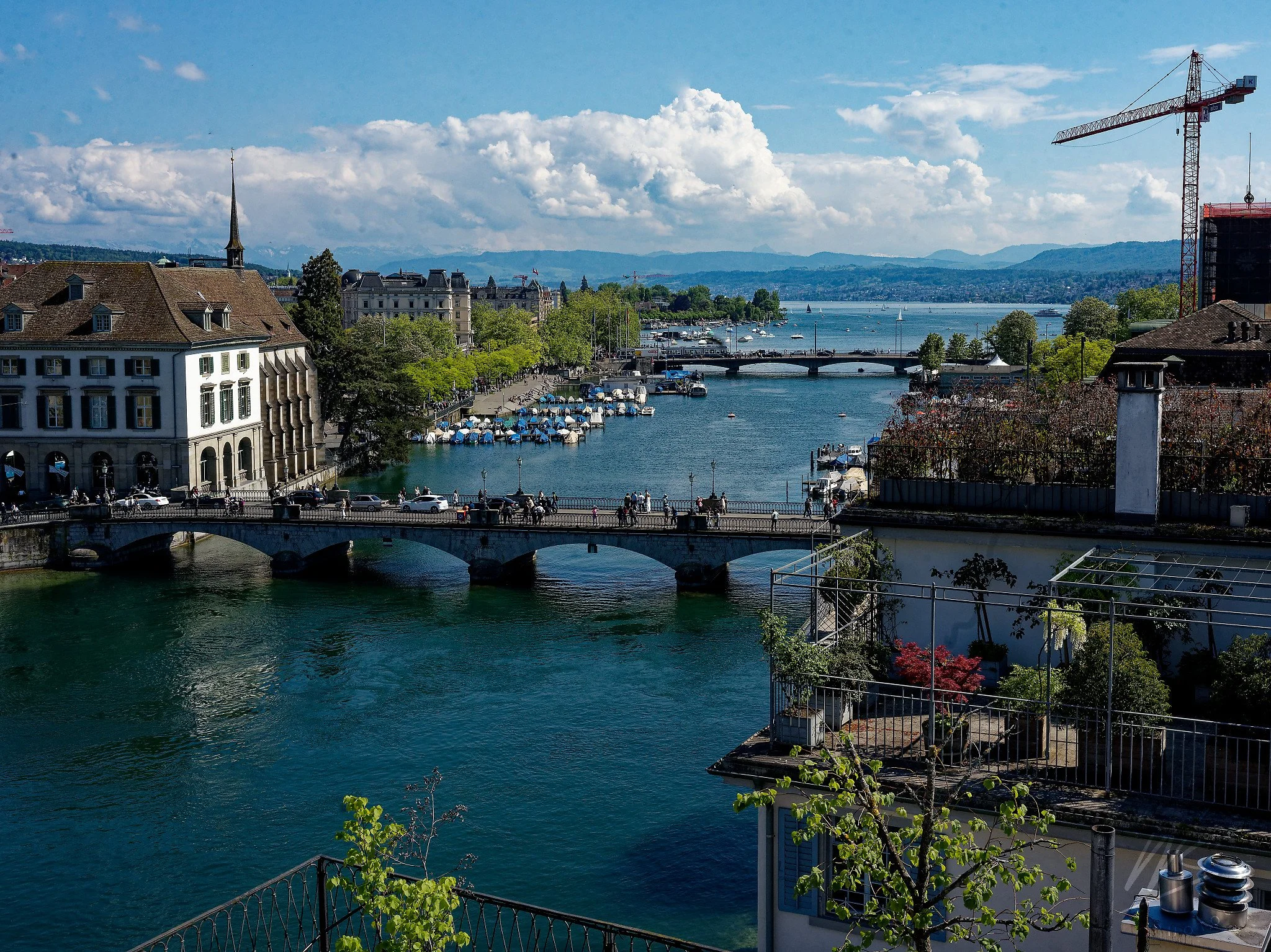 View south along the Limmat River toward Lake Zurich (Zürichsee). The Münsterbrücke crosses the foreground, with the Quaibrücke and the lakeside boat moorings of the Zürichsee promenade stretching beyond. The Alps are visible on the horizon on a clea