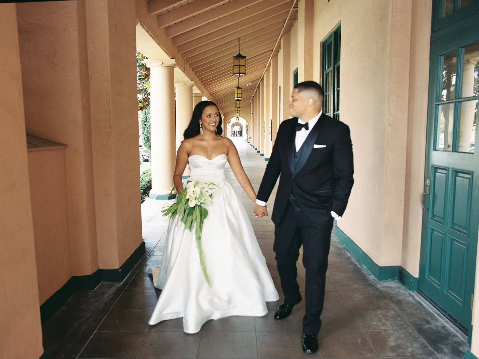 Bride and groom holding hands and walking in a corridor, smiling at each other. The bride wears a white strapless wedding gown with a bouquet, and the groom wears a black tuxedo with a bow tie.