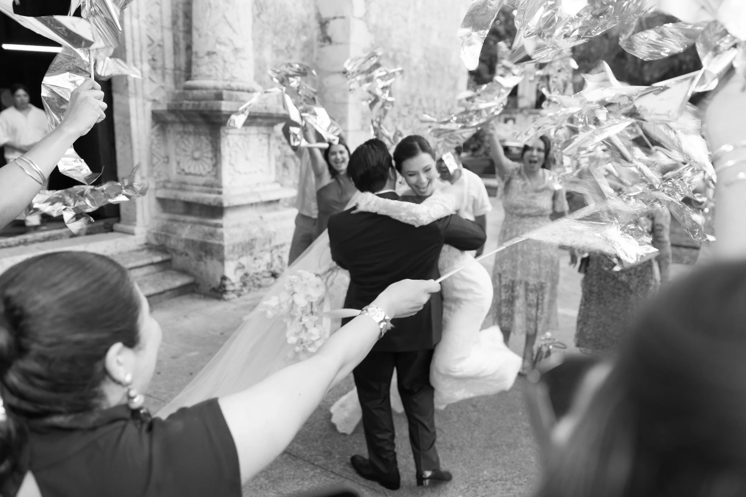 A bride and groom dance together surrounded by friends at a wedding celebration outside historic stone architecture. The bride is in a white gown, and the groom is in a dark suit, with friends holding shimmering decorations.