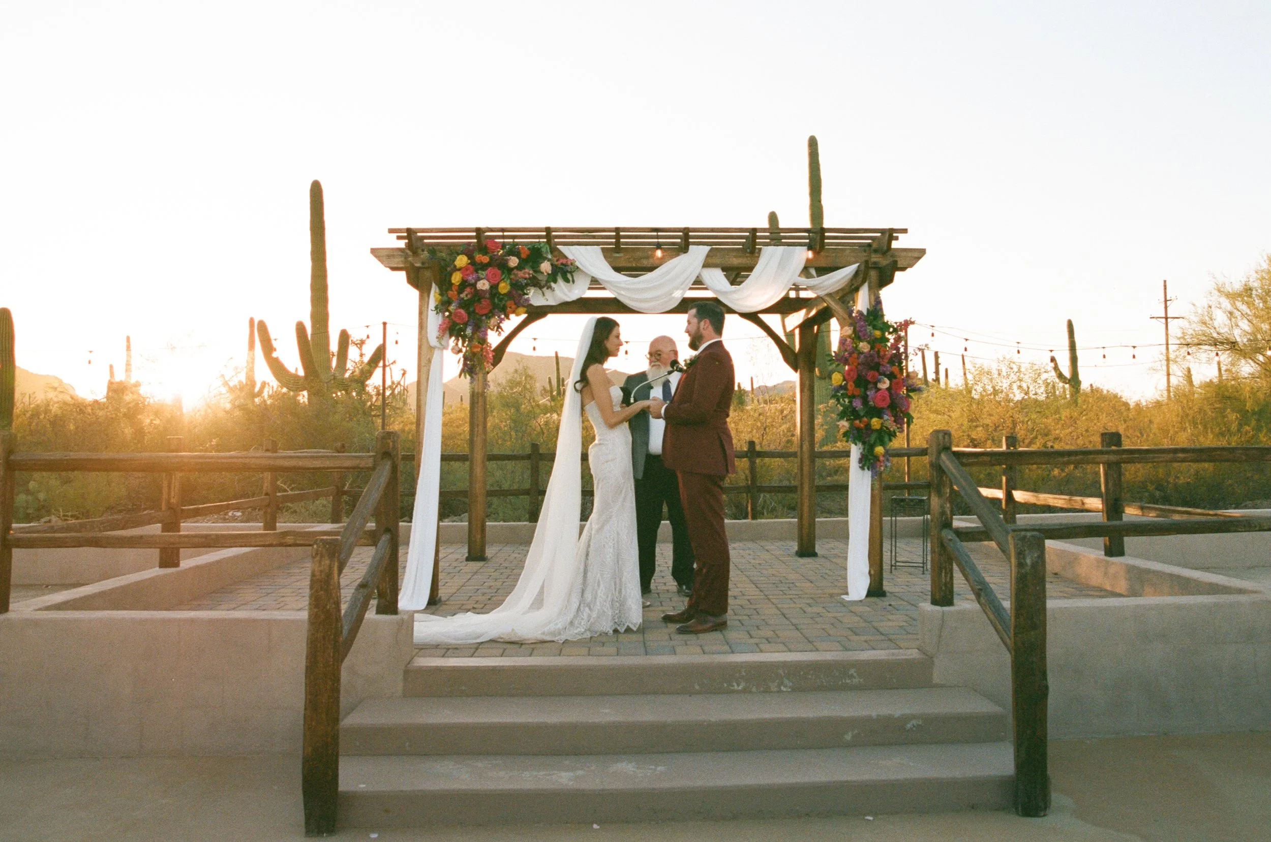 A couple getting married under a wooden gazebo decorated with white drapes and colorful flowers, outside during sunset with desert cacti in the background.