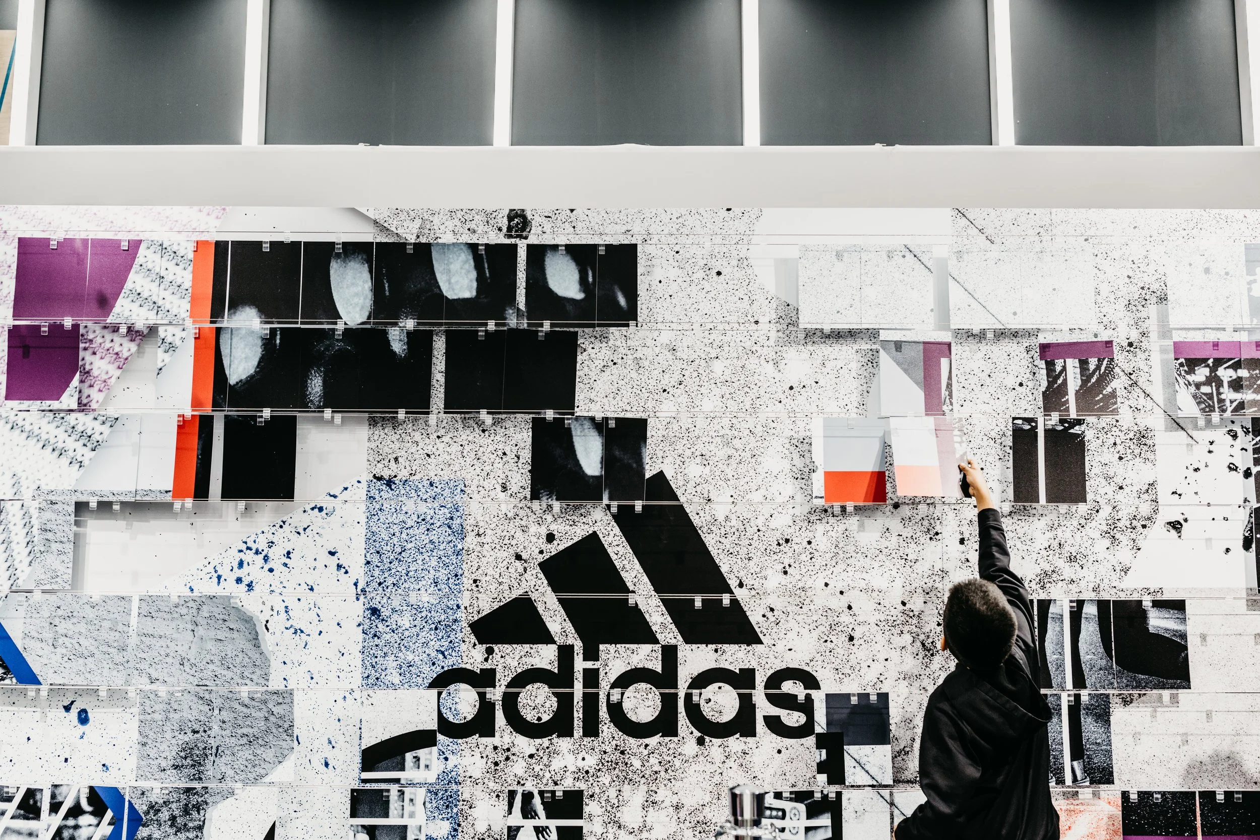 A person pointing at a large Adidas logo on a wall covered with various black, white, purple, red, and blue graphic designs.