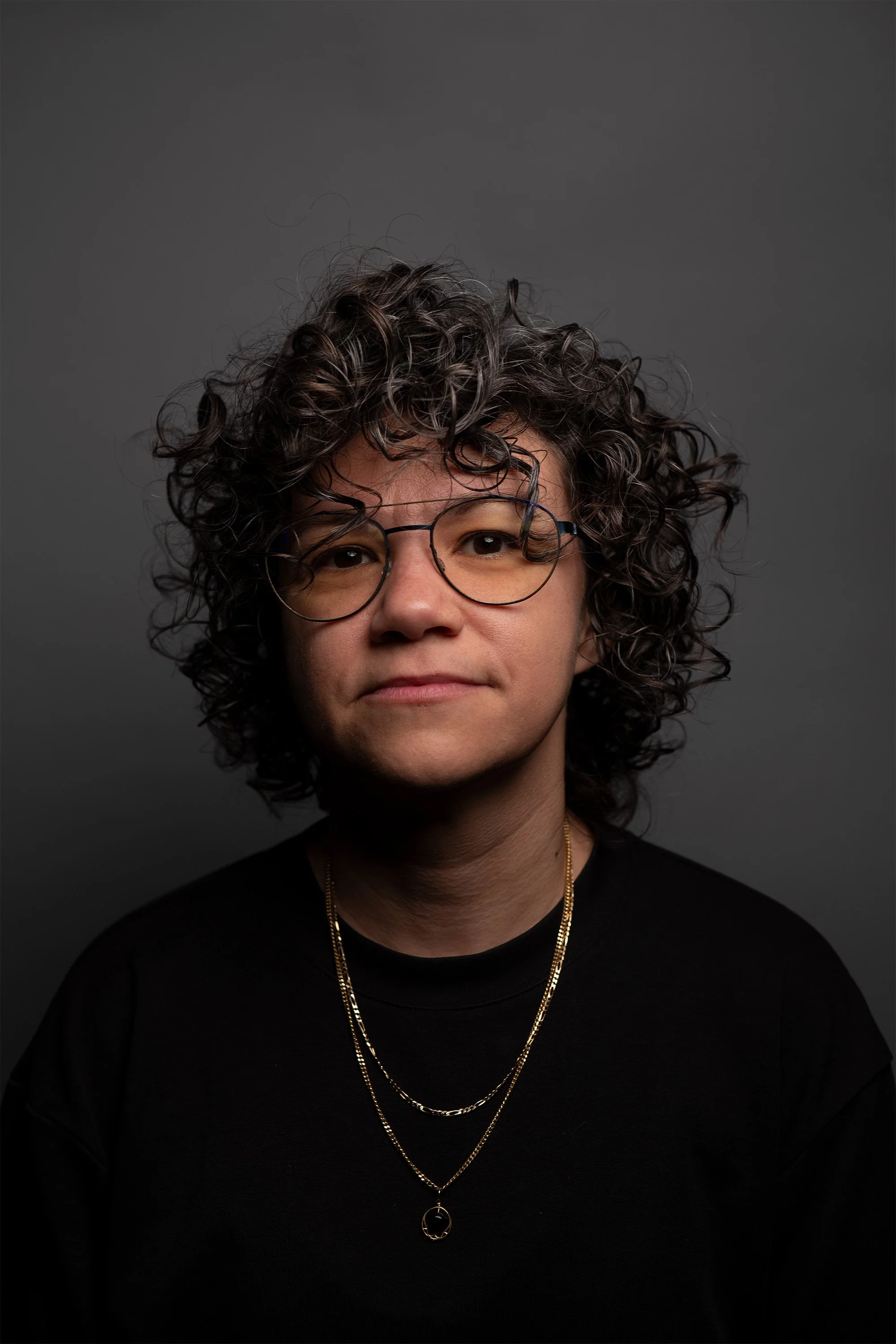 Portrait of a person with short, curly hair wearing glasses, a black top, and layered gold necklaces against a dark gray background.