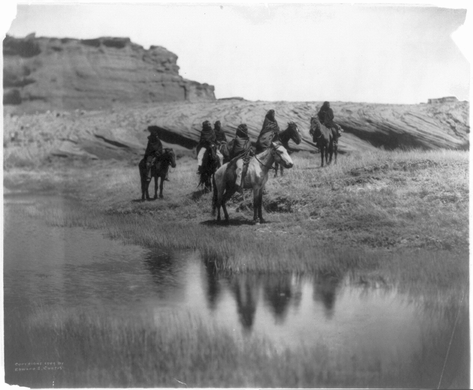 Six Navajo (Diné) riders wrapped in blankets sit on horseback at the edge of a still pool, their reflections in the water, with layered sandstone bluffs behind them. Photograph by Edward S. Curtis, 1904, Library of Congress.