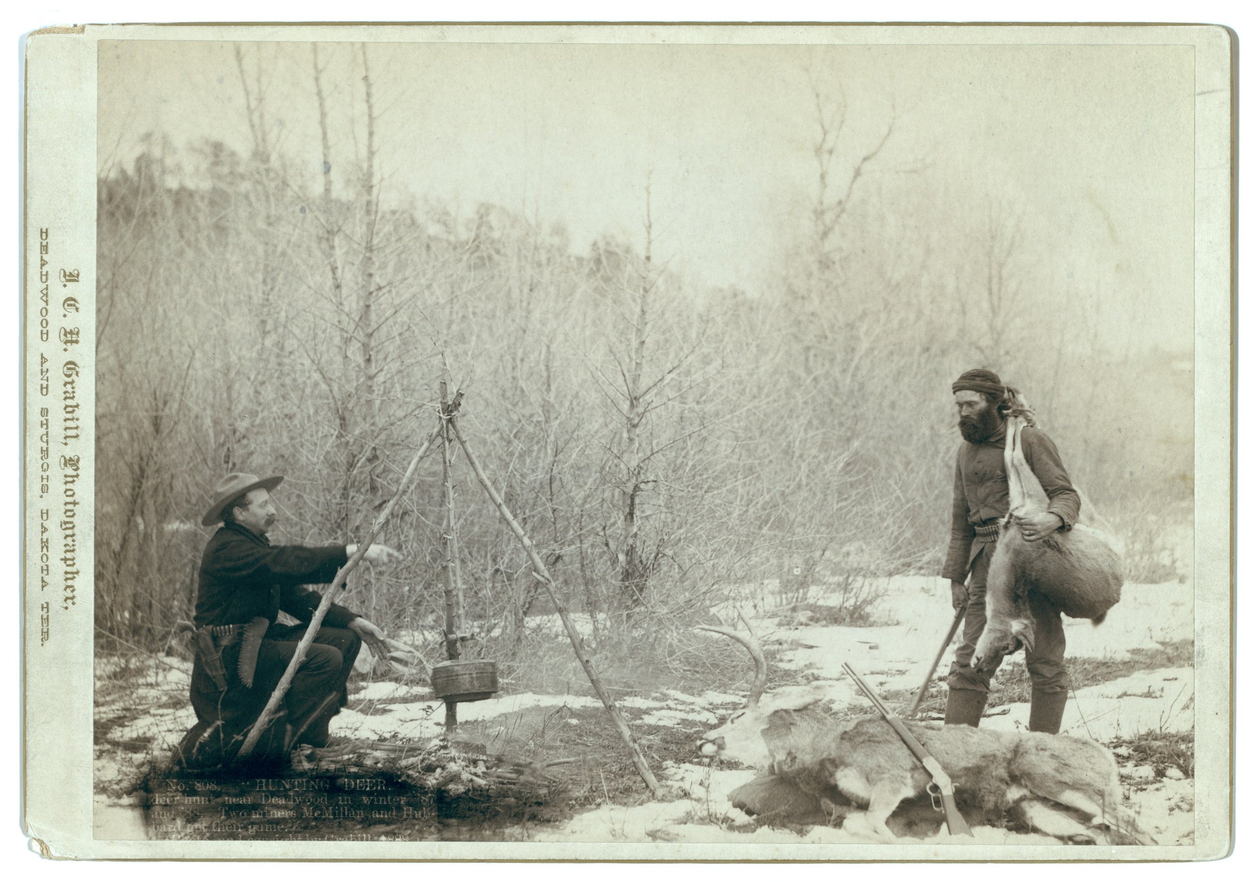 Two miners, McMillan and Hubbard, at a snowy winter camp near Deadwood, Dakota Territory, 1887–88, one cooking over a tripod fire, the other carrying a deer on his shoulder. Photograph by John C. H. Grabill, Library of Congress.
