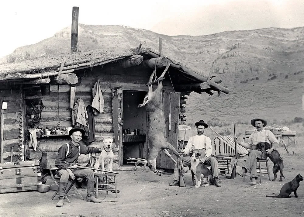 Three hunters sit with their rifles and dogs outside a sod-roofed log cabin on the Williams Fork in northwestern Colorado, circa 1899, with a deer hanging from the eaves and a black cat in the foreground.