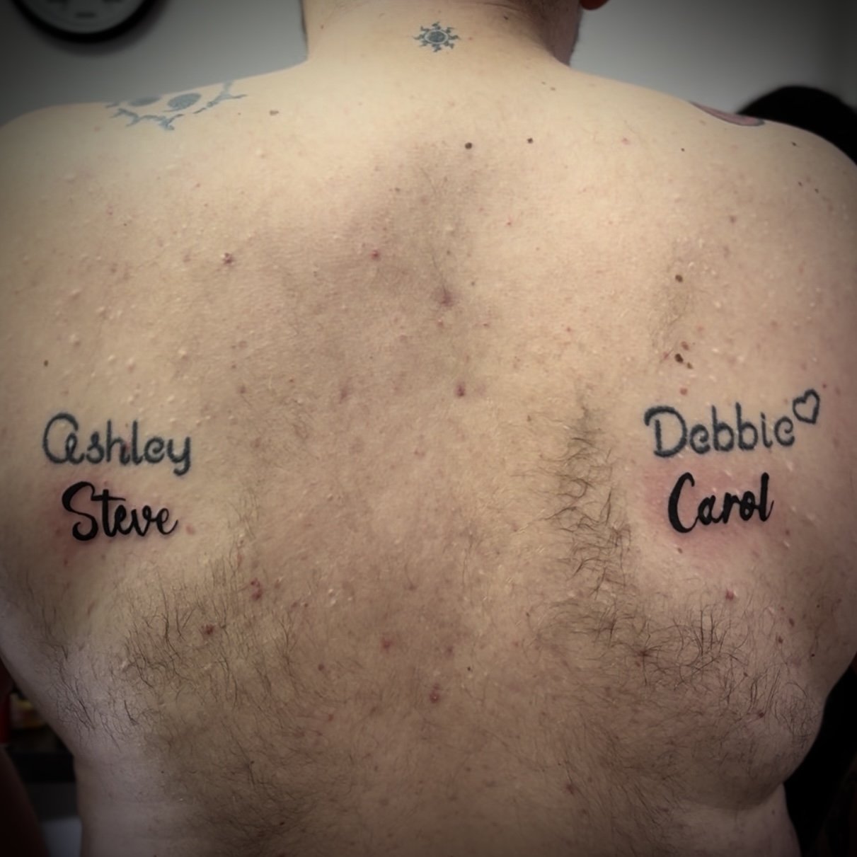 Rear view of a person's back with tattoos featuring names "Ashley" and "Steve" on the left shoulder, and "Debbie" with a heart and "Carol" on the right shoulder.