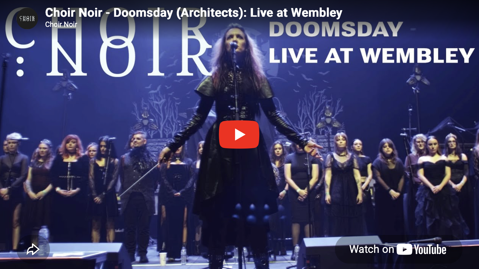 A female choir conductor leading a choir on stage during a live performance at Wembley, with the words 'Choir Noir - Doomsday (Architects): Live at Wembley' displayed in the background.