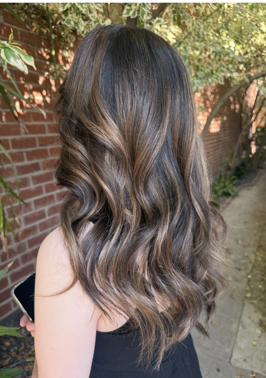 Woman with long, wavy brown hair with lighter highlights, standing outdoors near a brick wall and green foliage.