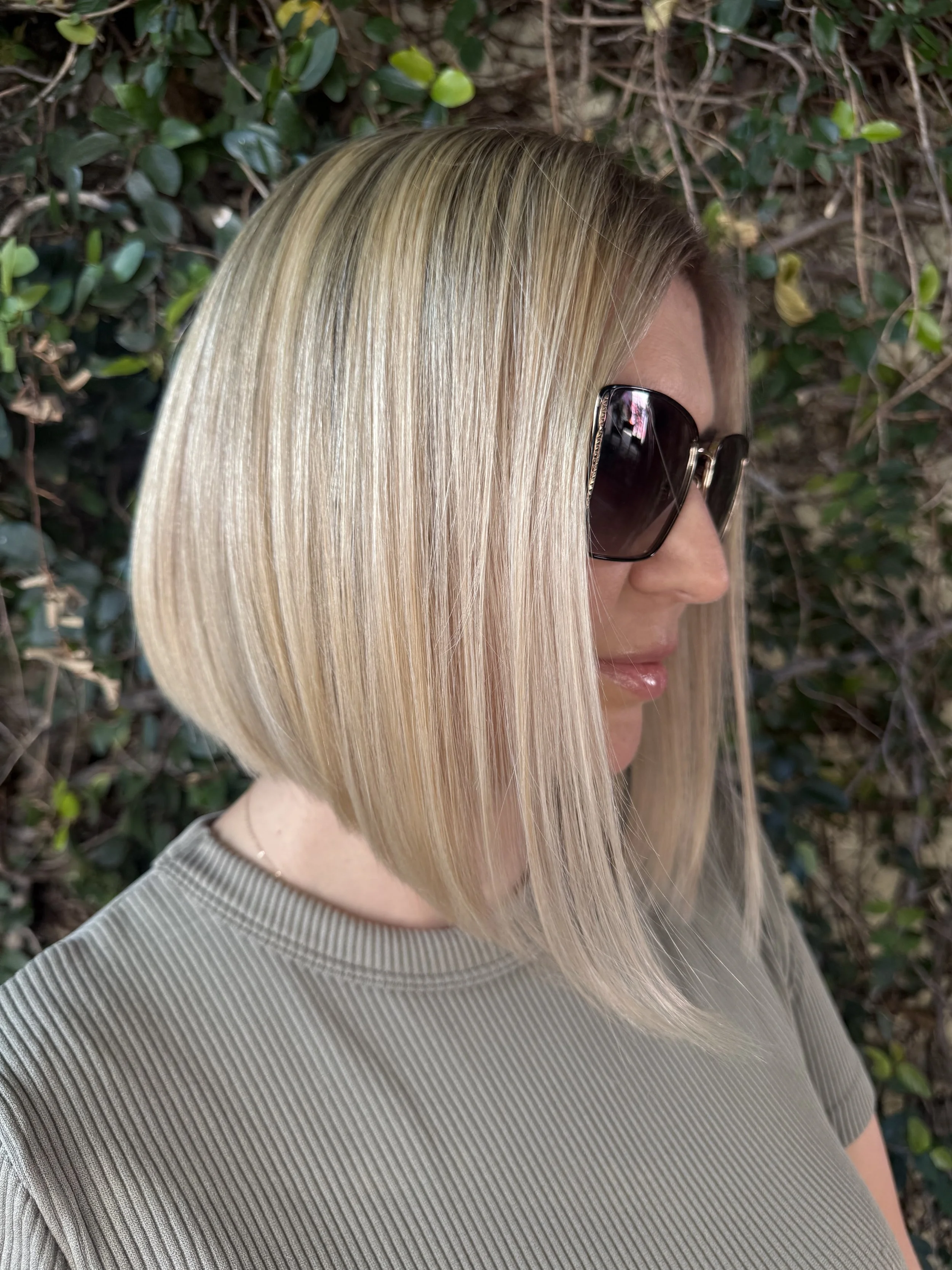 Side profile of a woman with long, straight, blonde hair wearing large sunglasses and a ribbed gray shirt, standing outdoors near green foliage.