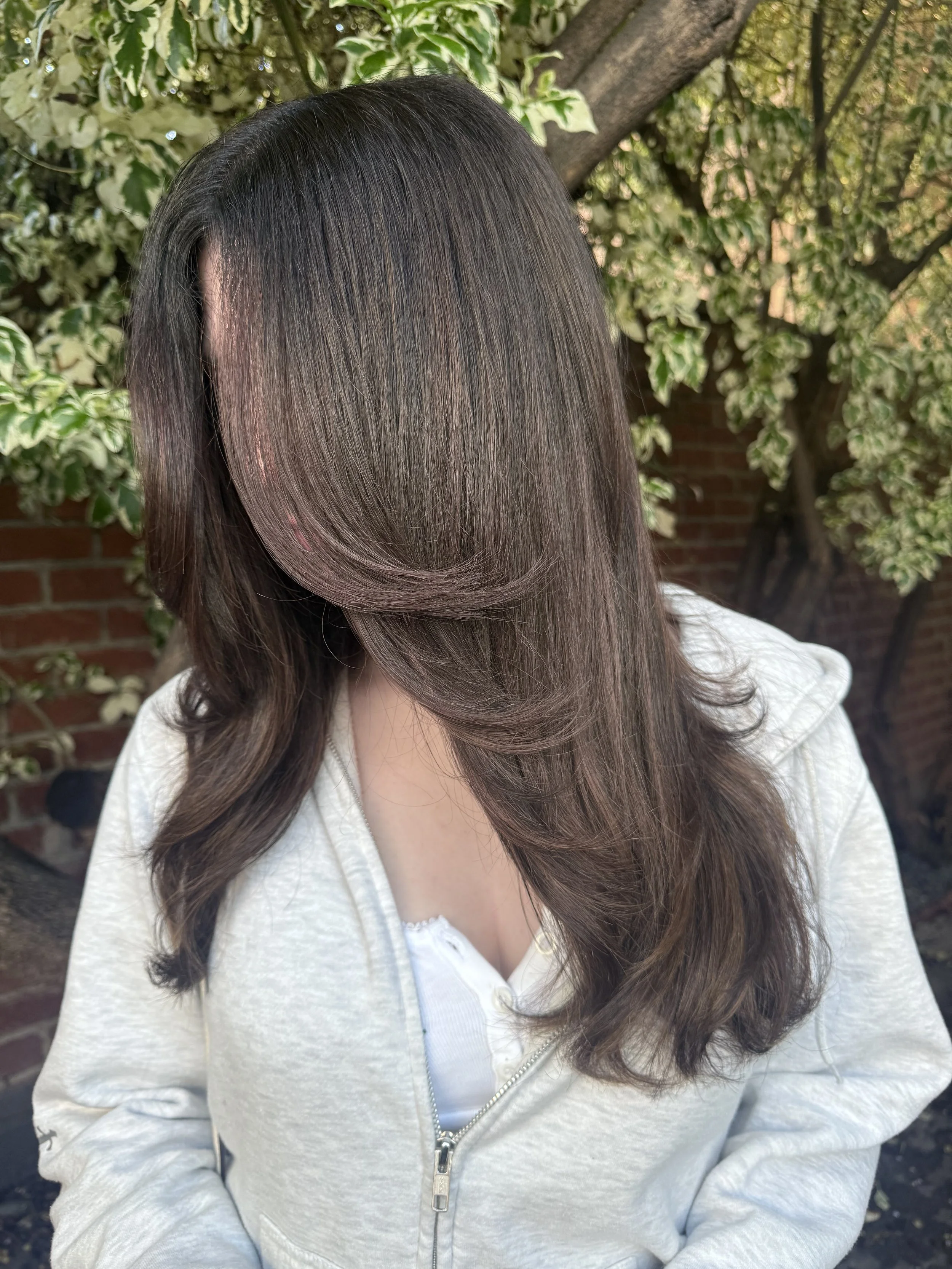 A woman with long, dark brown hair with subtle highlights, wearing a white zip-up sweater, sitting outdoors near a tree with green and cream variegated leaves and a brick wall in the background.