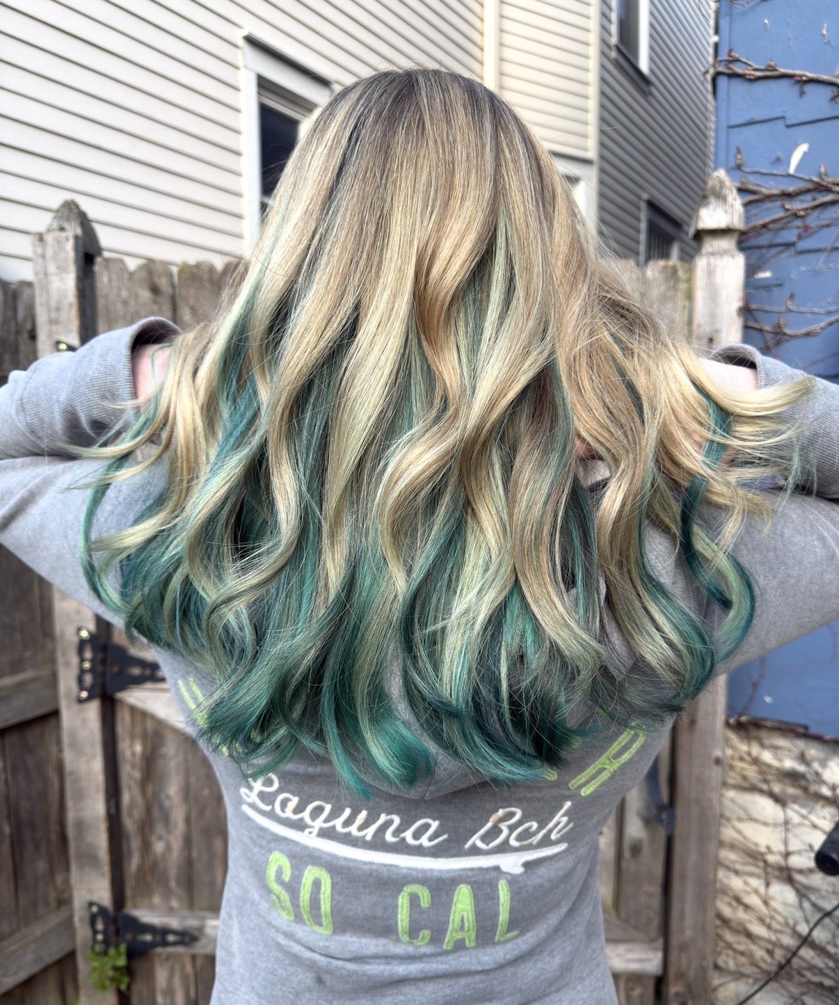 Back view of a woman with long, wavy blonde hair with green ombre tips, standing outdoors near a wooden fence and house.
