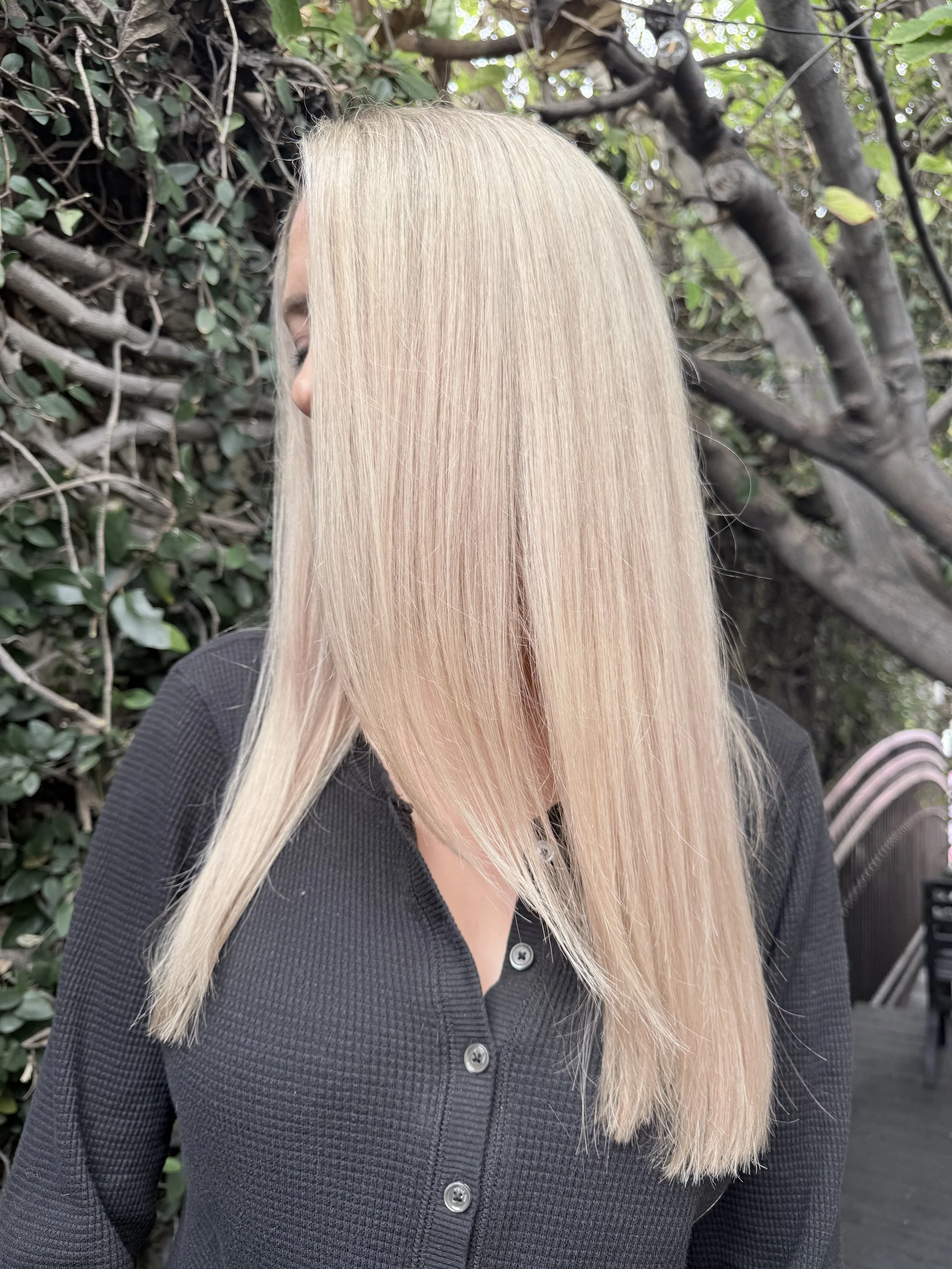A woman with long, straight blonde hair wearing a black button-up shirt stands outdoors with green foliage and tree branches in the background.