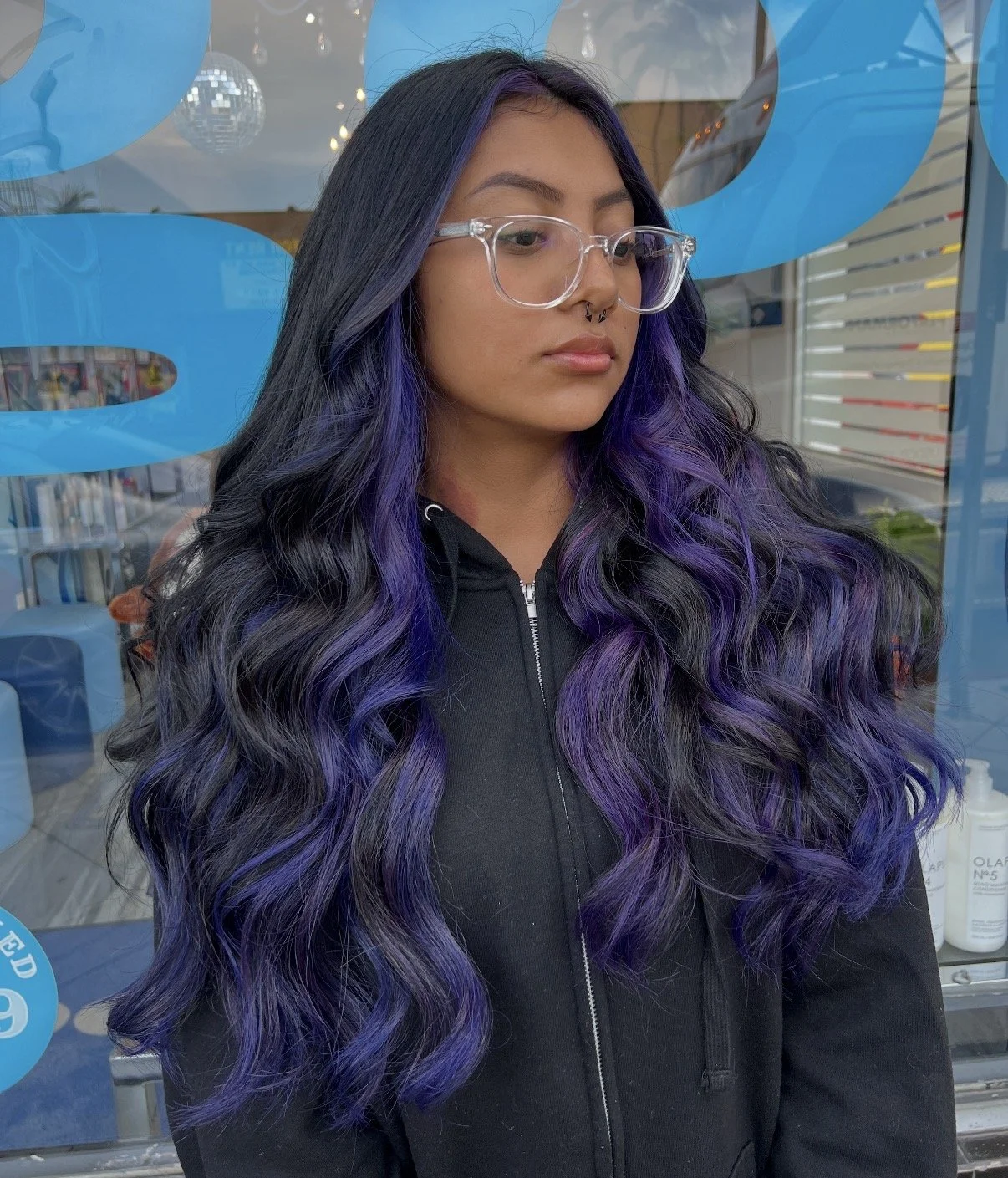 Young woman with long wavy black and purple hair, wearing clear glasses and a black zip-up jacket, standing outside near a glass window with blue design elements and reflection of a parking lot.