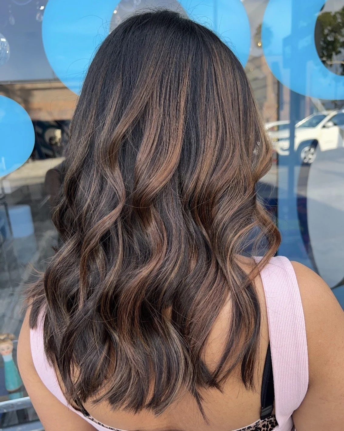 The image shows a woman with long, wavy hair dyed with a blend of dark brown and light caramel highlights, wearing a pink tank top, standing outside in front of a glass window that reflects cars and a street scene.
