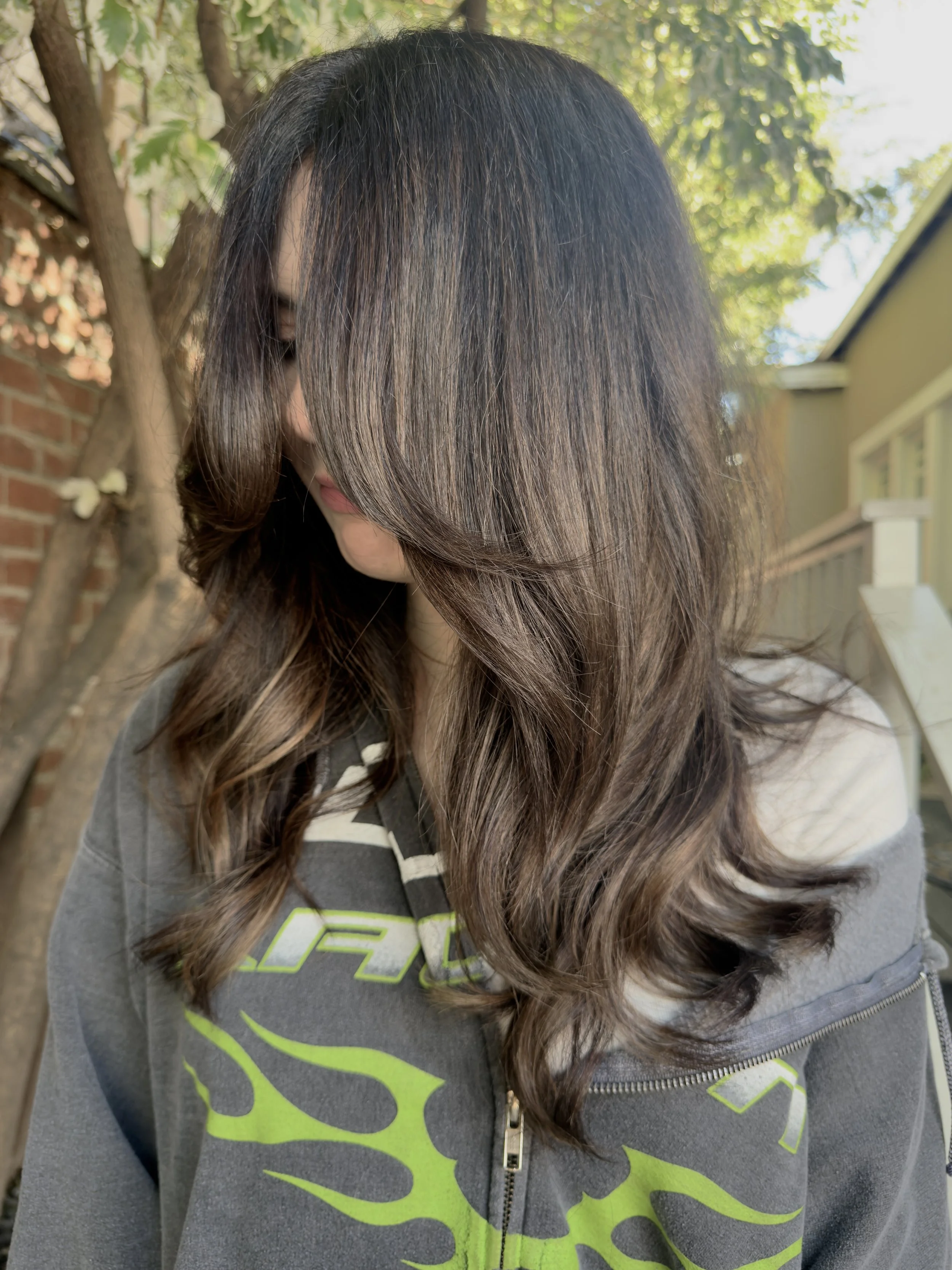 A person with dark brown, wavy hair partially covering their face, wearing a gray hoodie with neon green graphic design, outside with green foliage and house in the background.