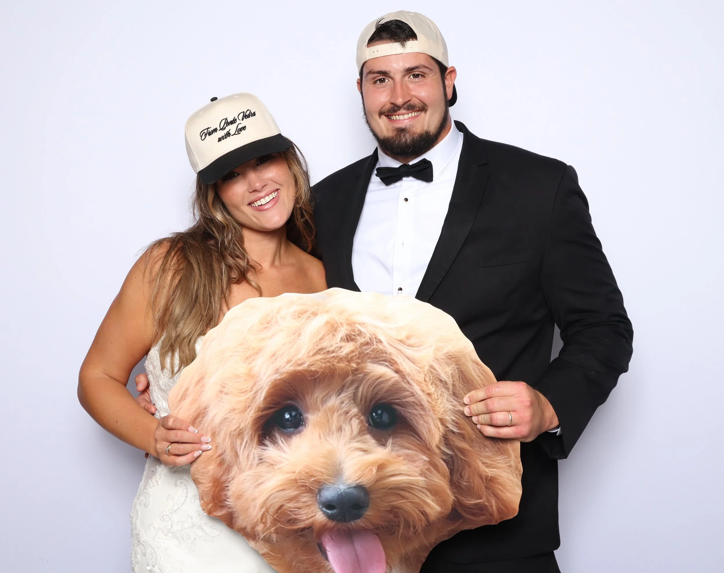 A smiling woman in a white dress with a cap that says 'From Dusk Till Dawn with Love' and a man in a tuxedo with a bow tie, both holding a large photo of a fluffy puppy with big eyes and tongue sticking out against a plain background.
