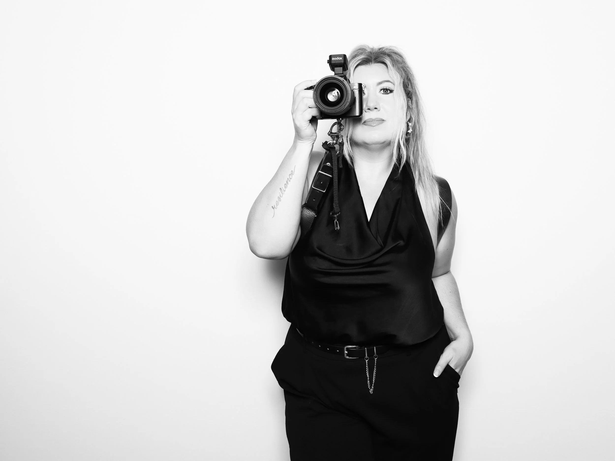 A woman with blonde hair holding a camera to her eye, wearing a black sleeveless top and black pants, standing against a plain white background.