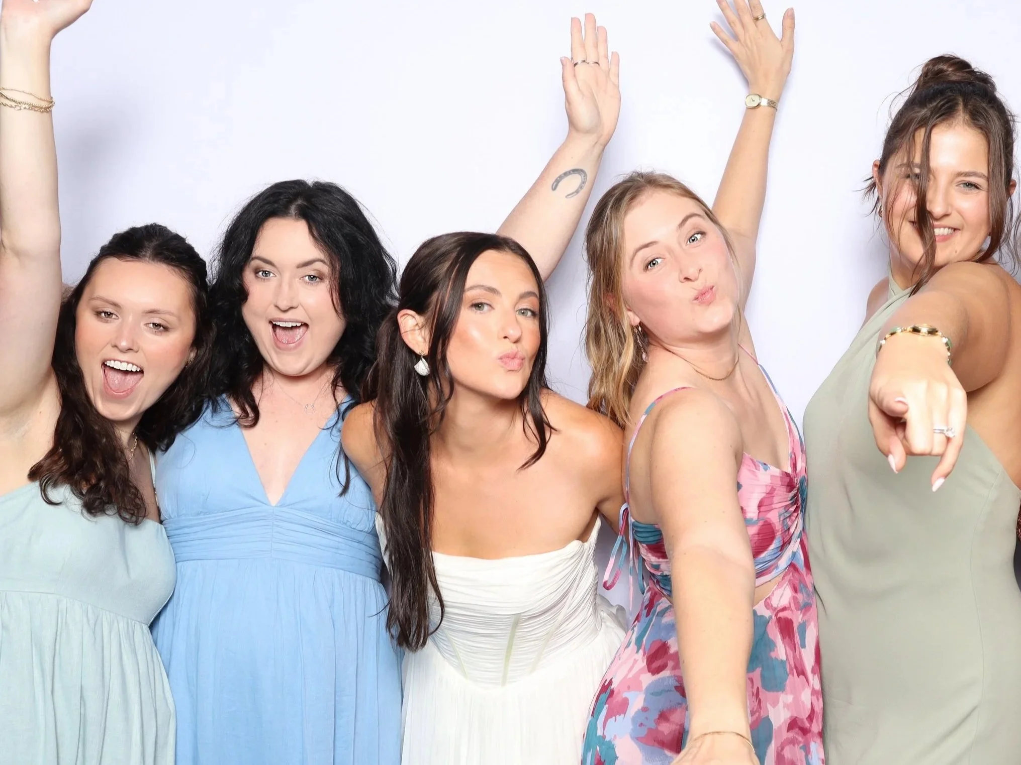 Group of five women celebrating, smiling, and posing for the camera with their arms raised and pointing, against a plain white background.