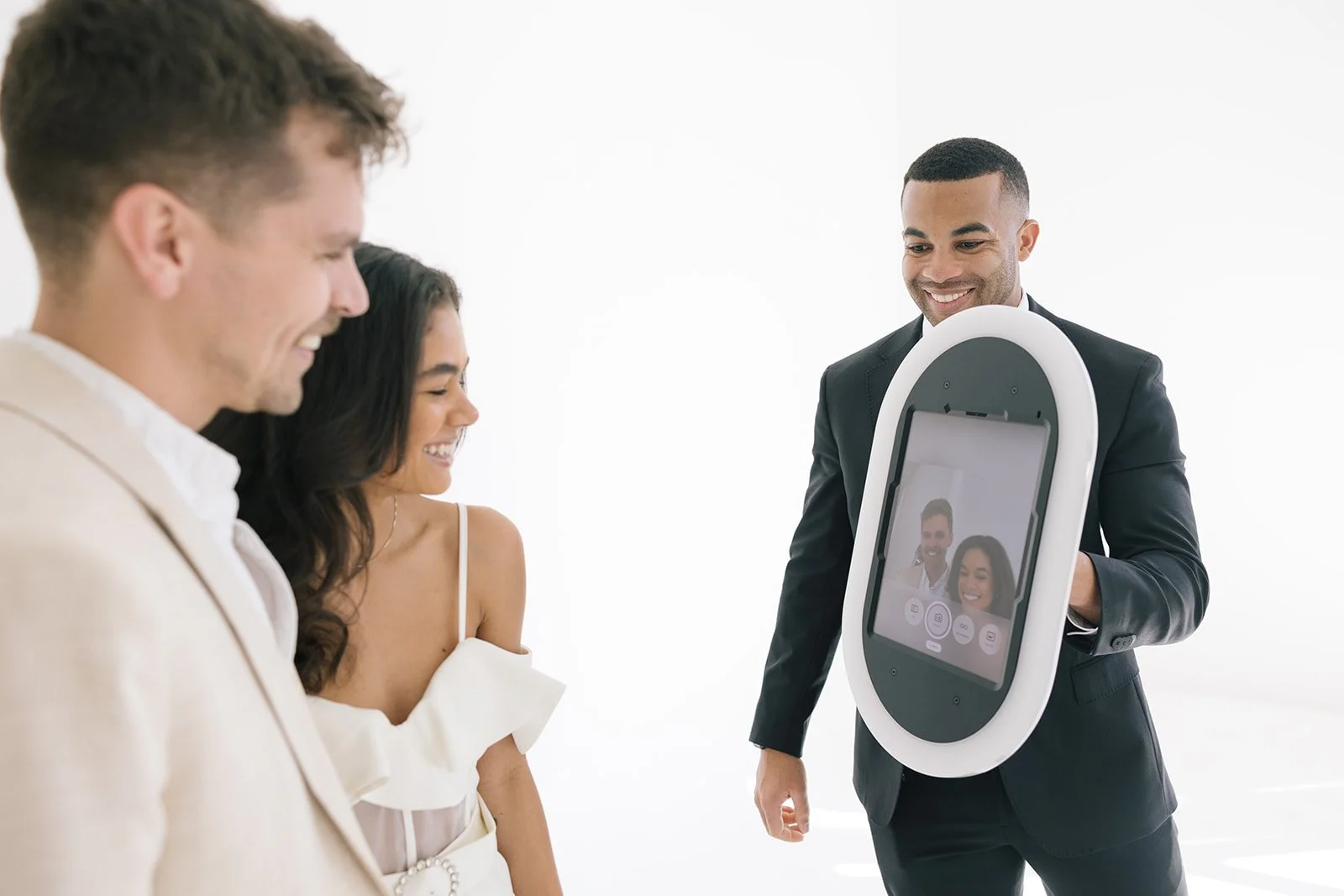 A man in a black suit holding a device with a screen showing a photo of two smiling people, with two women and one man in front of him smiling and looking at the device.