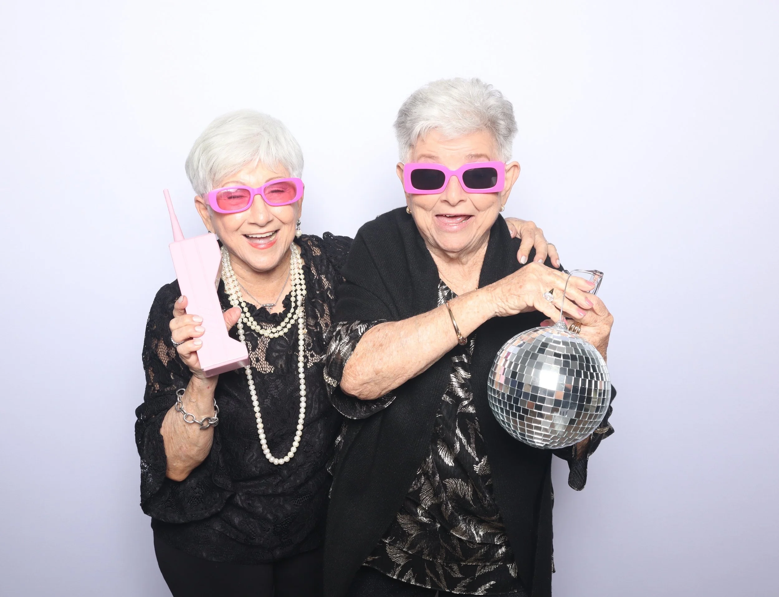 Two elderly women celebrating, one holding a pink vintage telephone and the other holding a disco ball, both wearing pink sunglasses, smiling, and dressed in black with accessories, against a plain white background.