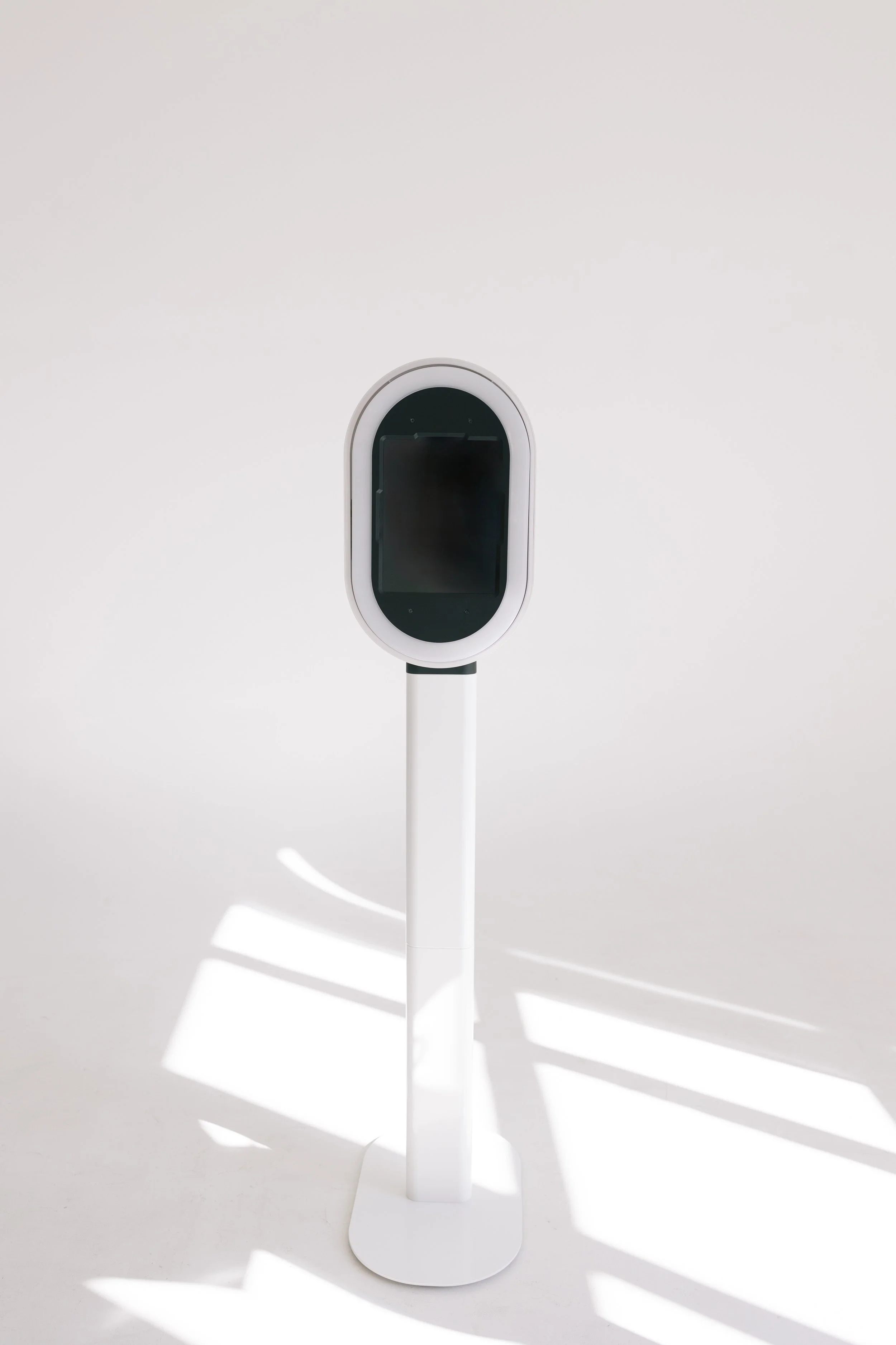 A sleek, white, modern stand with a black screen on top, standing upright on a plain white background with shadows cast by sunlight.