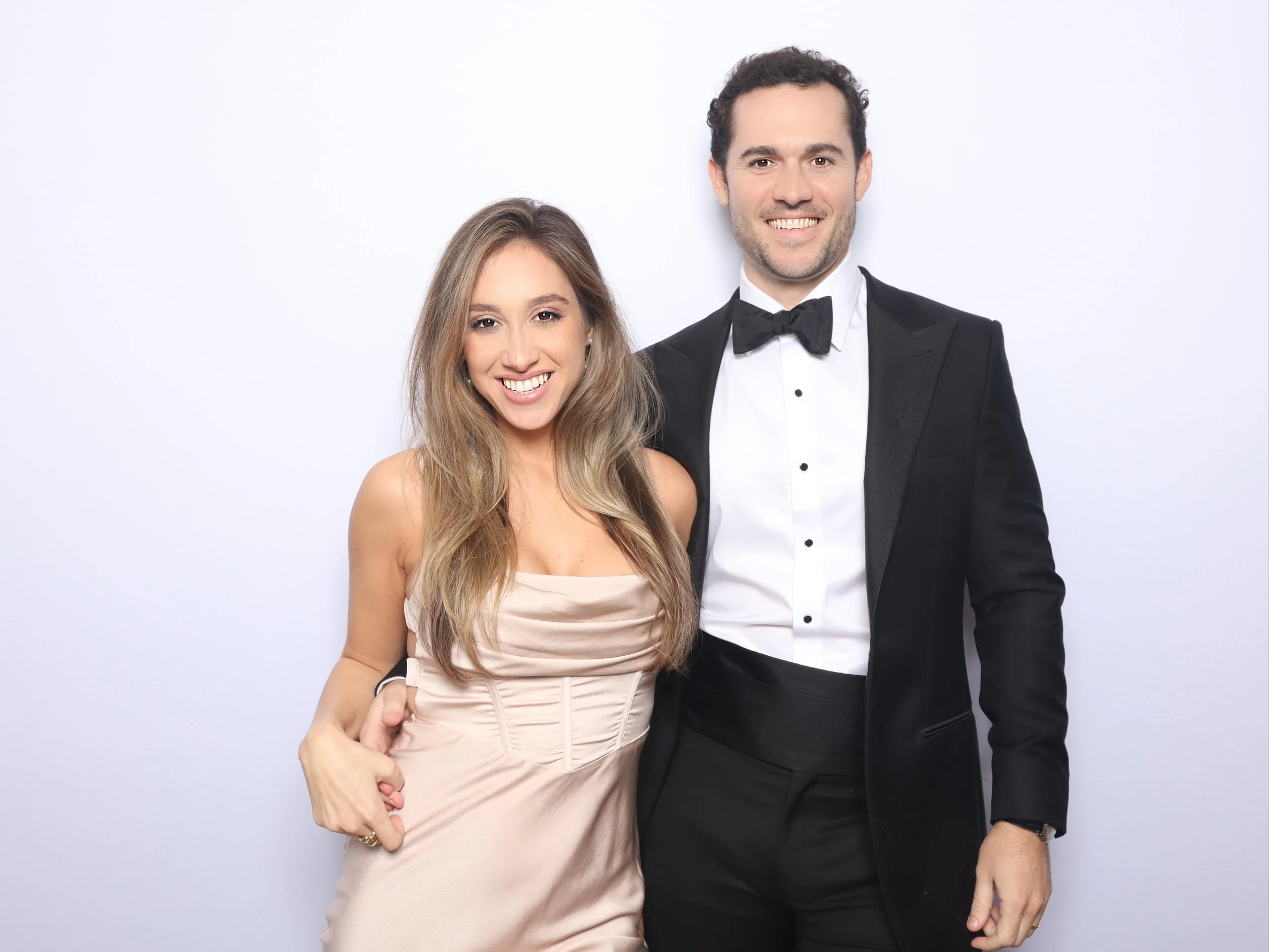 A man in a tuxedo and a woman in a strapless dress posing together, both smiling, against a plain white background.