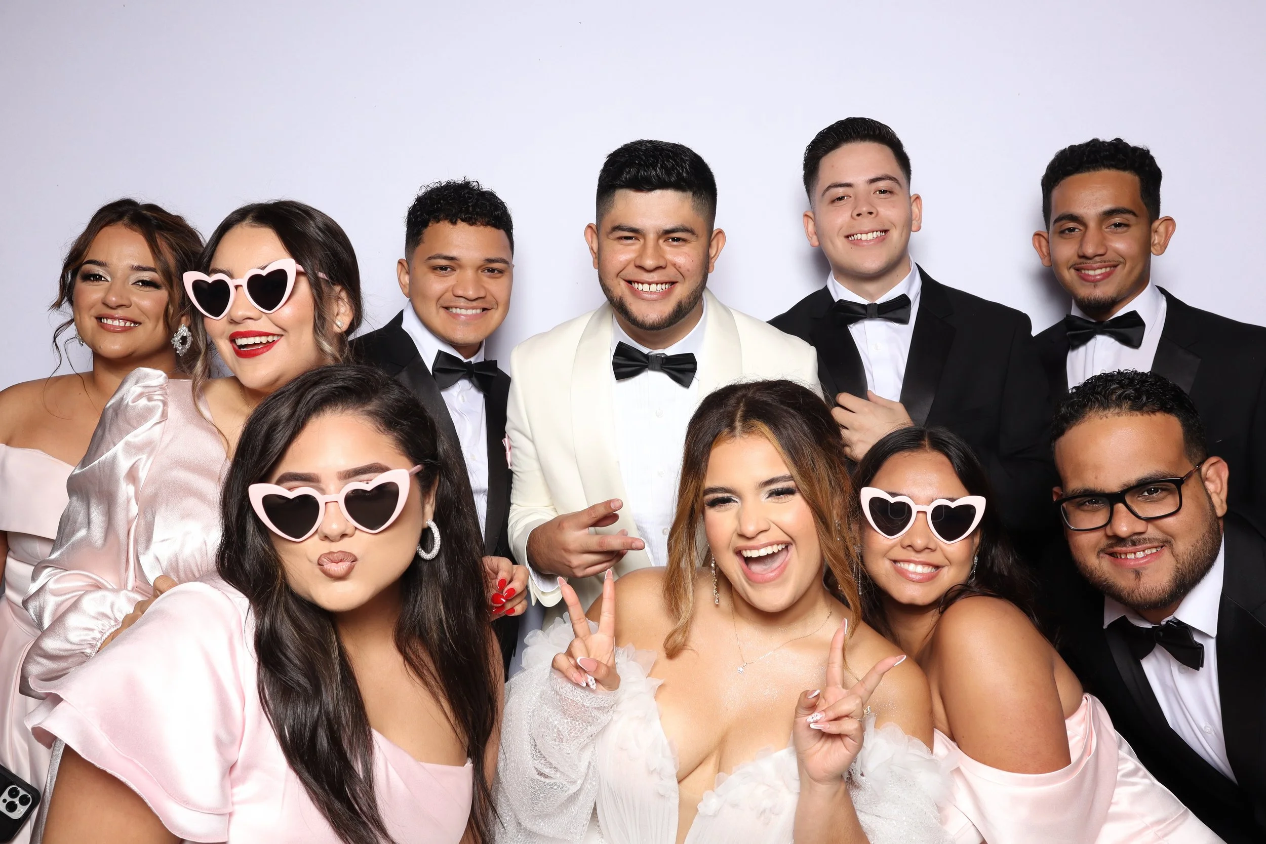 Group of young adults dressed in formal attire, smiling and making playful gestures, some wearing sunglasses, in front of a plain white background.