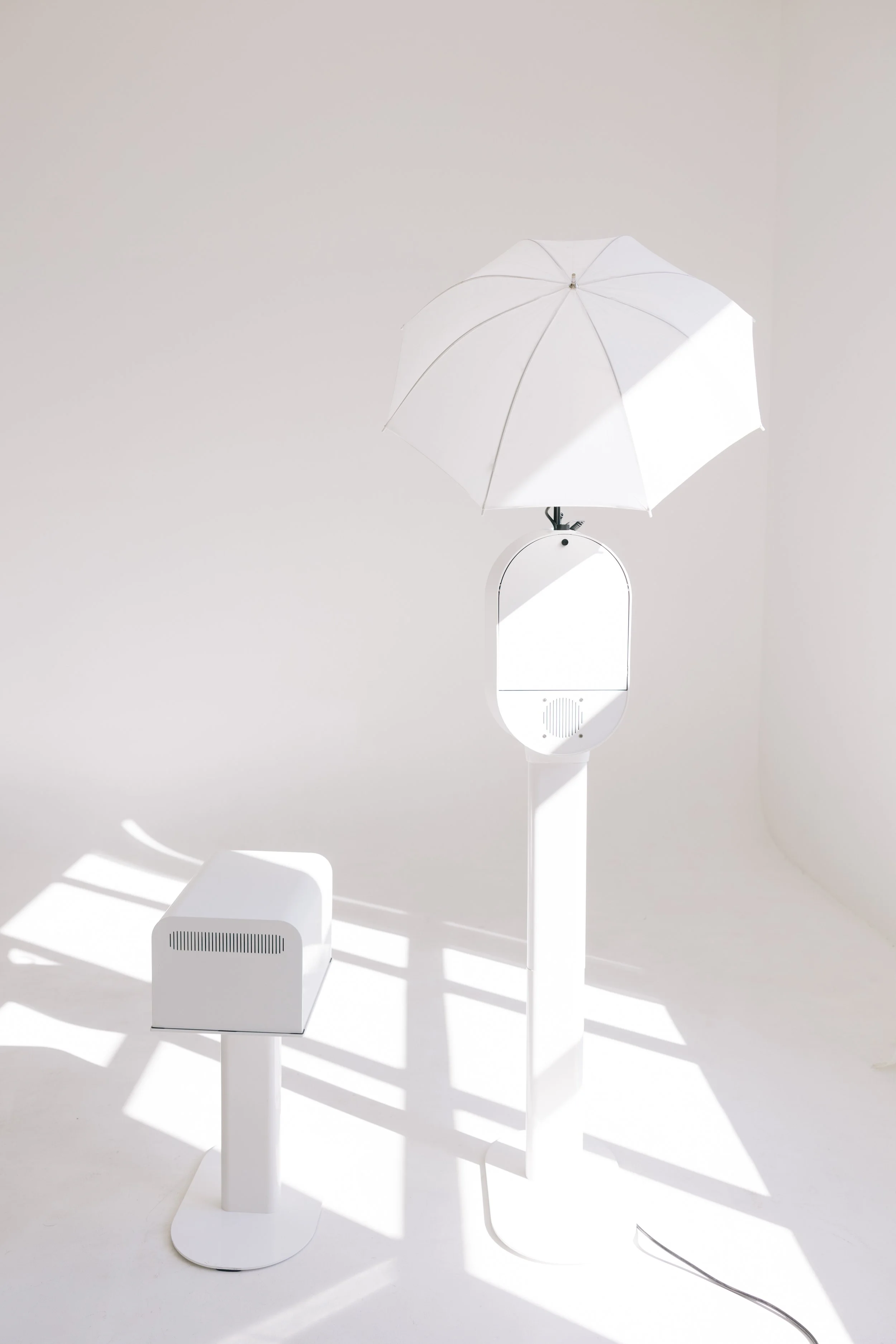 White device with an umbrella on top, resembling a modern, minimalist art installation with shadows cast on the floor.